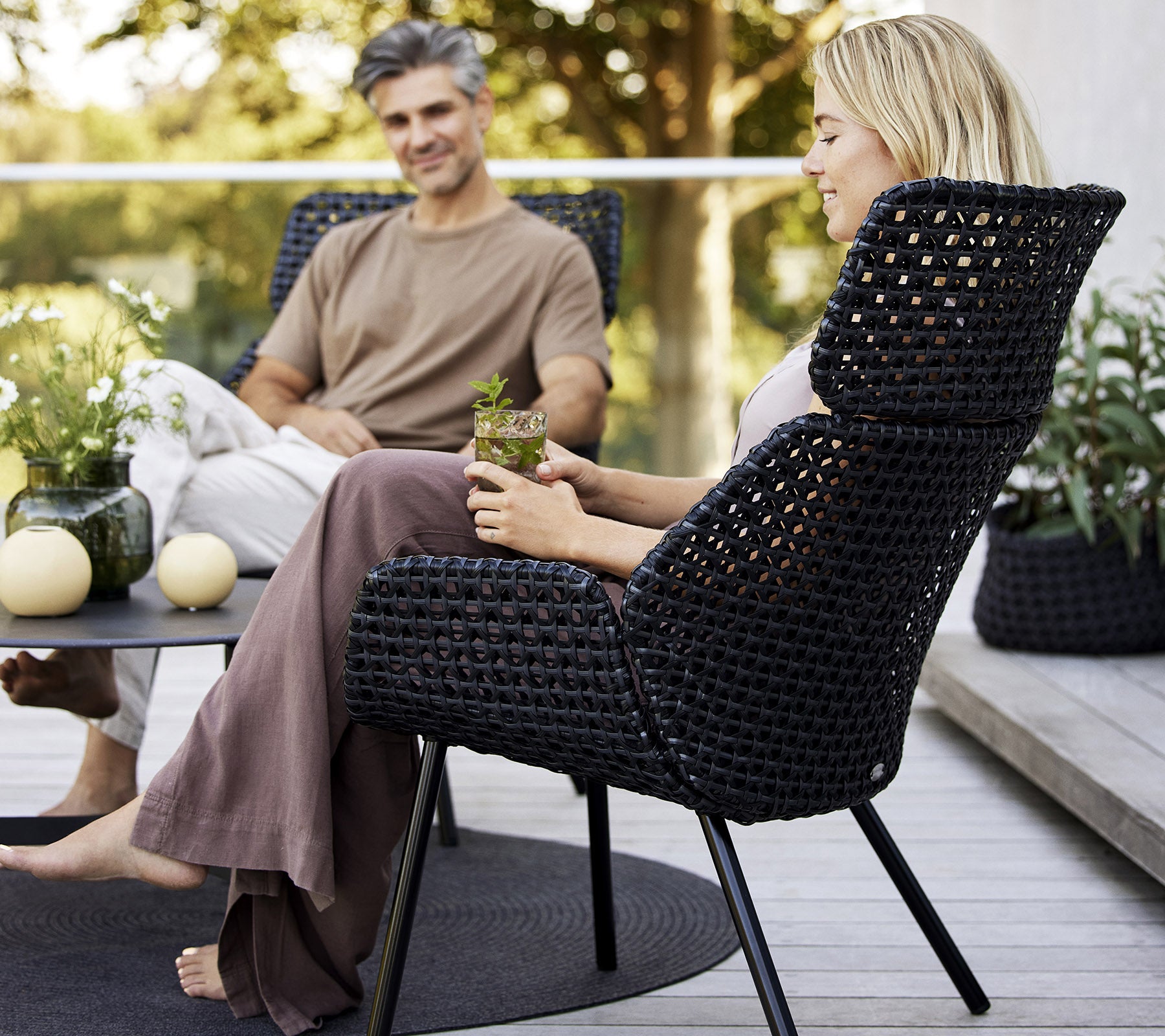 A stylish highback chair in dark woven material, featuring two people enjoying a relaxed moment with plants and decor around them.