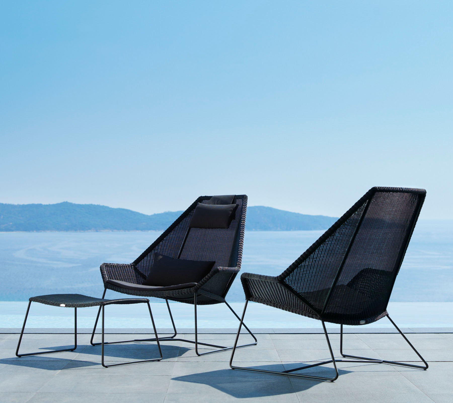 Two stylish, modern chairs with sleek lines and open mesh design, accompanied by a matching footrest, overlooking a serene water view.