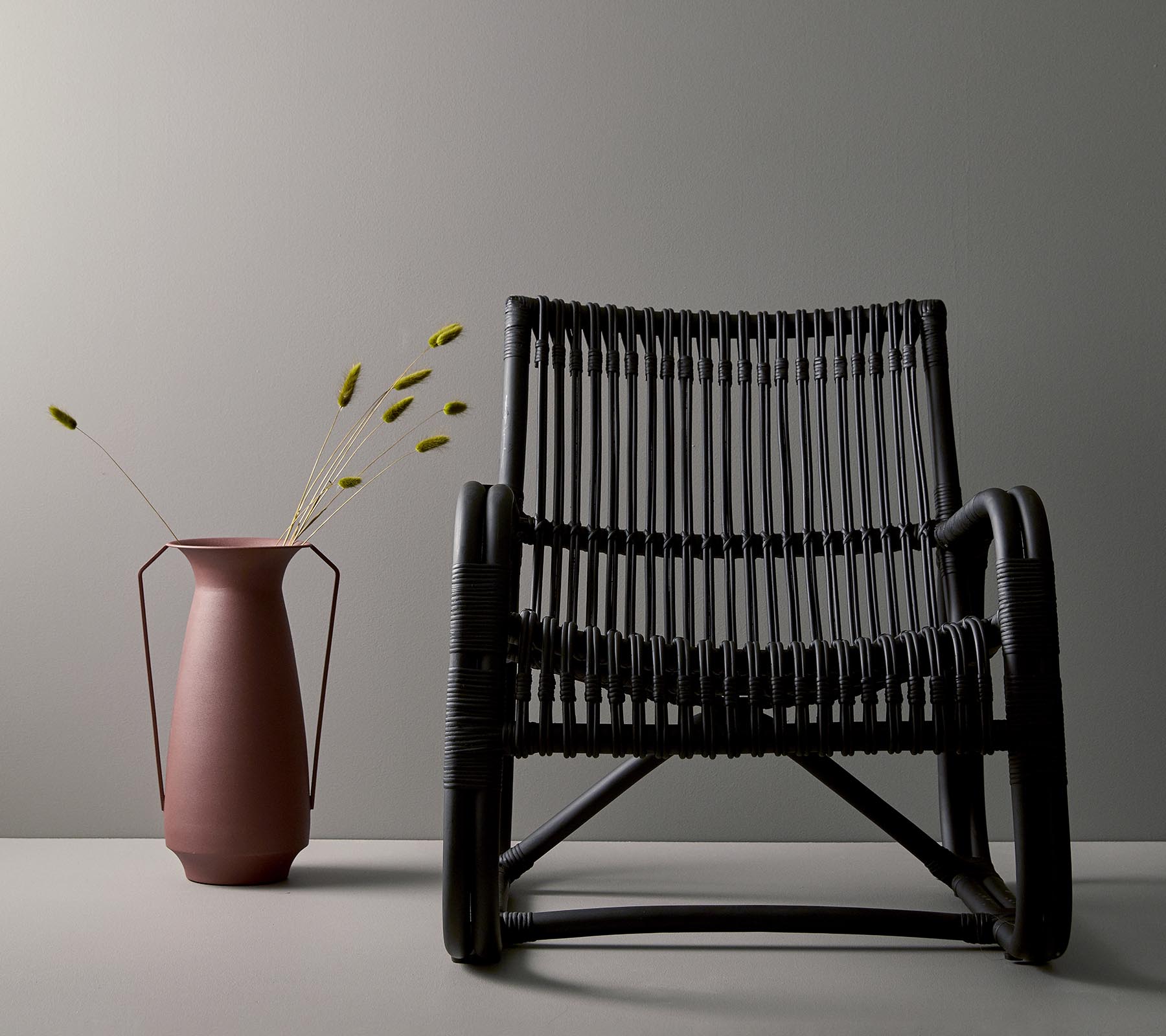 Stylish black woven chair next to a pink vase with green stems, set against a minimalist gray background.