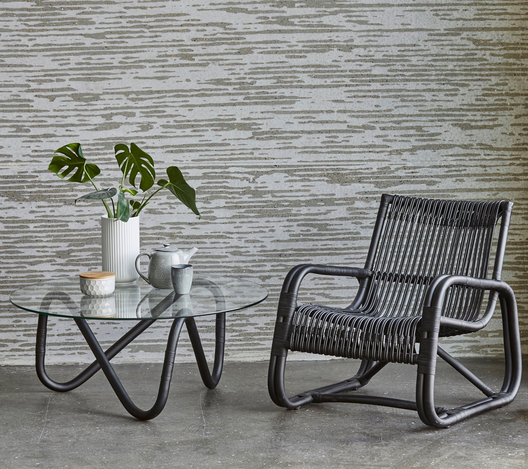 A stylish chair and a glass-top table with a plant and coffee set, complemented by a textured wall background.