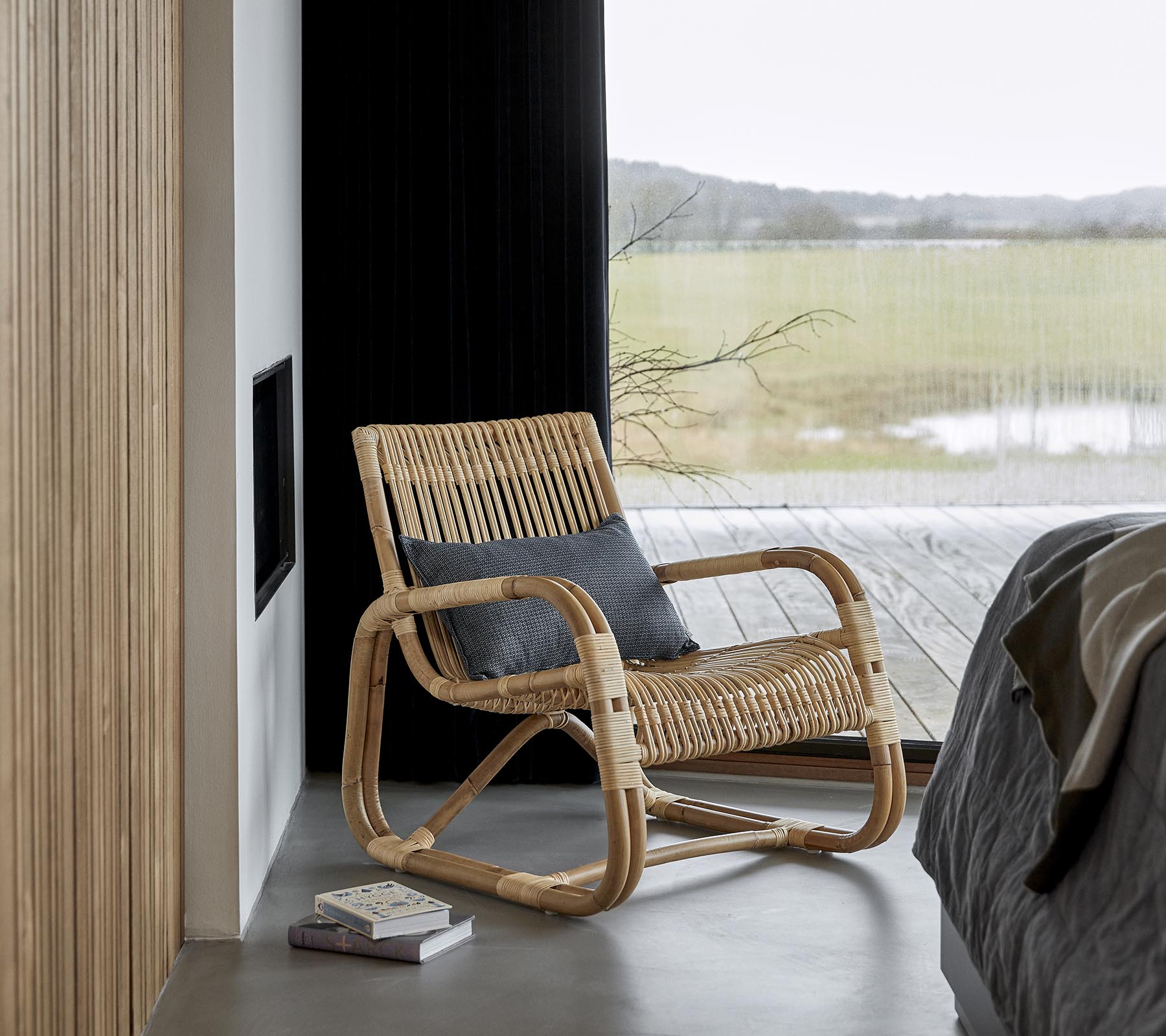 A stylish rocking chair made of natural materials, placed next to a bed and a book, with a scenic view visible through large windows.