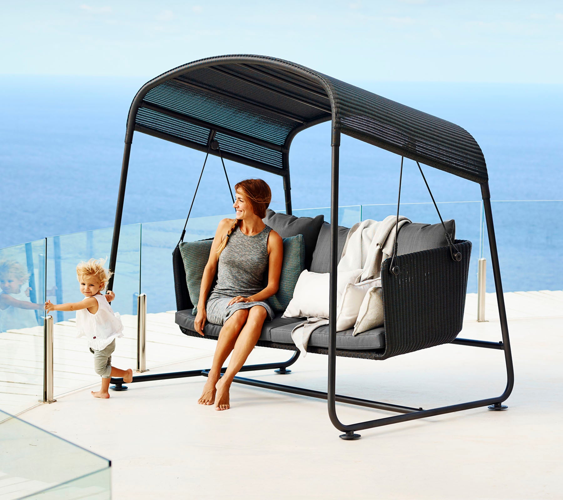 A stylish swing seat with comfortable cushions, featuring a woman relaxing and a child playing nearby, set against a serene ocean backdrop.