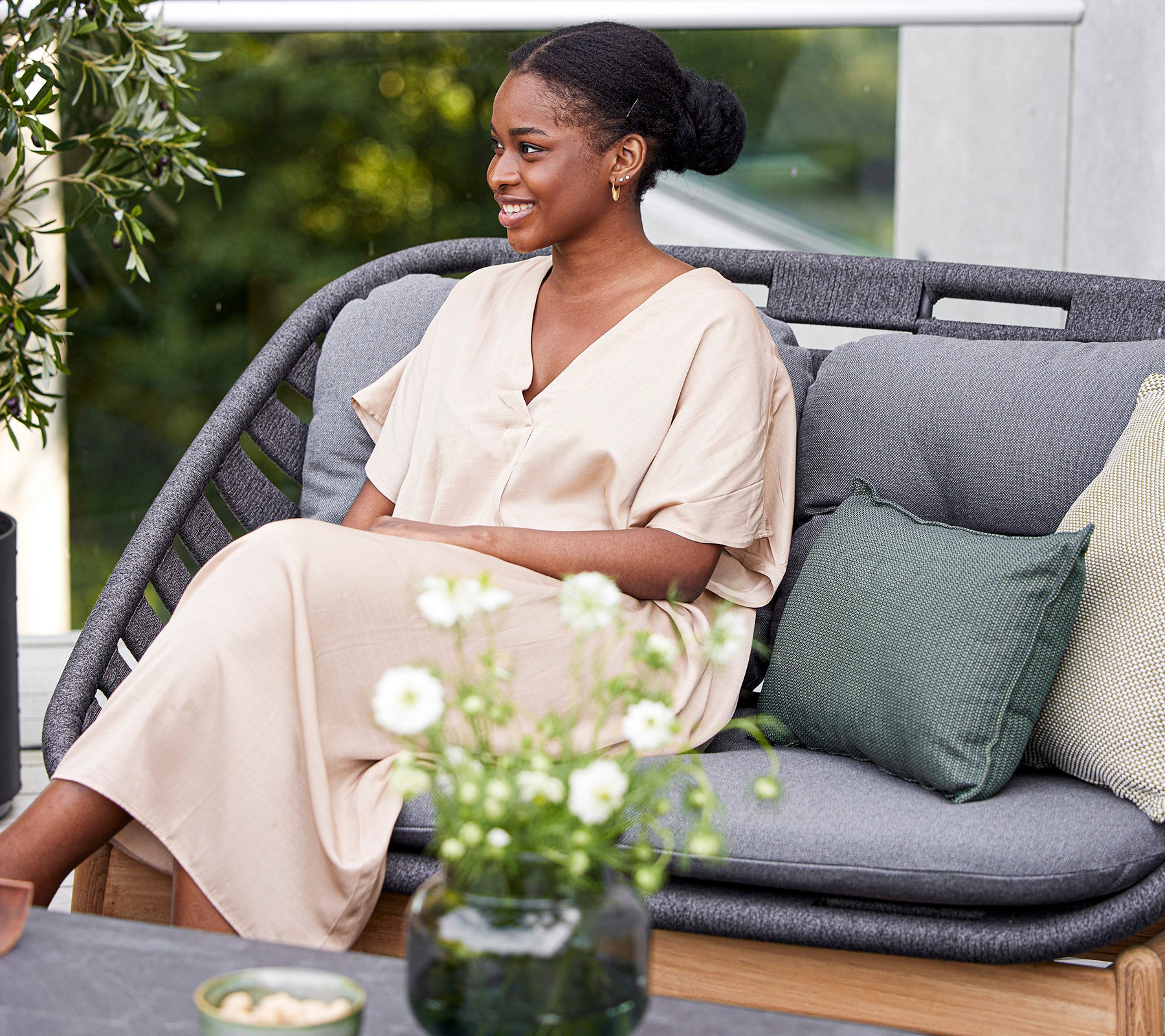 A smiling woman in a light-colored dress sits on a stylish sofa, surrounded by decorative cushions and a small floral arrangement.