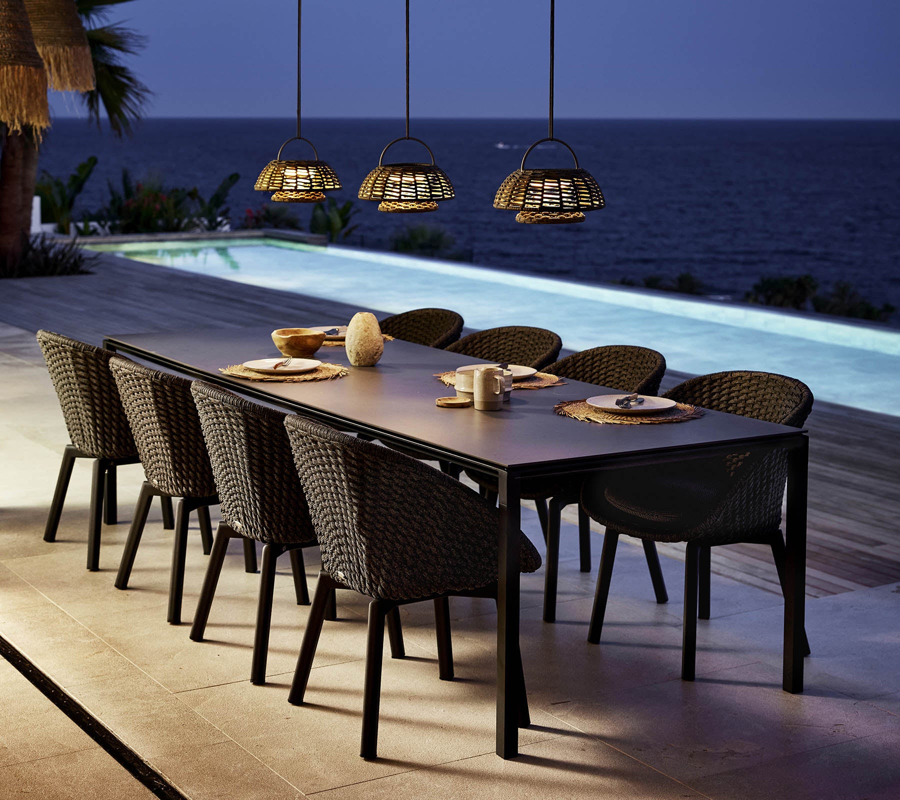 A stylish dining setup features a long table surrounded by peacock chairs, illuminated by pendant lights above a serene water view.