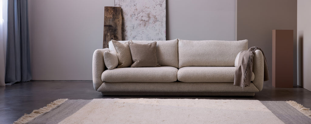 Modern beige sofa with pillows and a grey throw in a minimalist lounge setting.