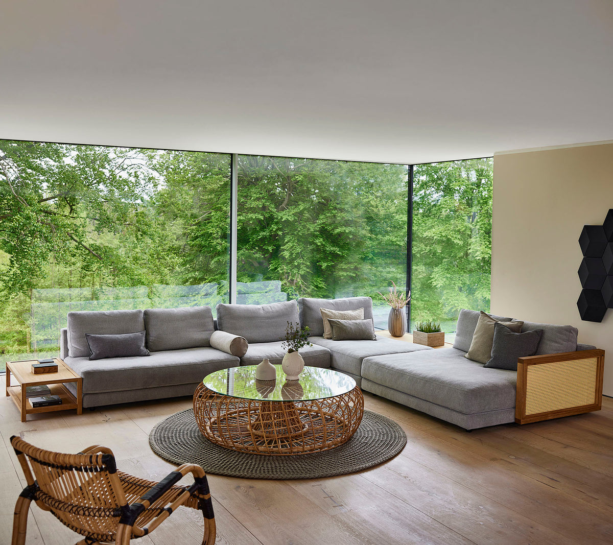A modern seating area featuring a gray sectional sofa, a round coffee table, and a woven chair, surrounded by greenery.