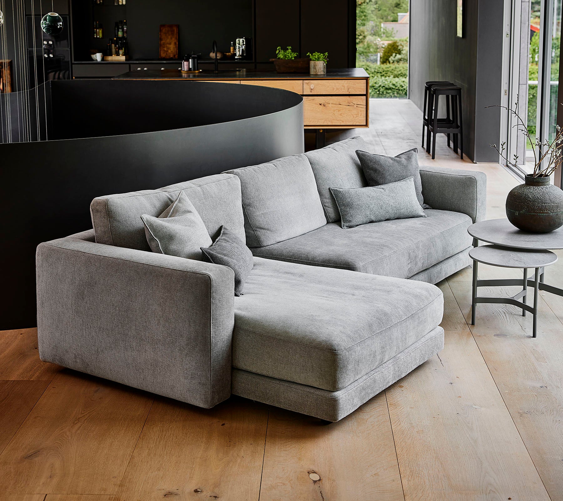 Modern grey sectional sofa with multiple pillows, accompanied by a round coffee table and decorative vase. Simple, elegant design.