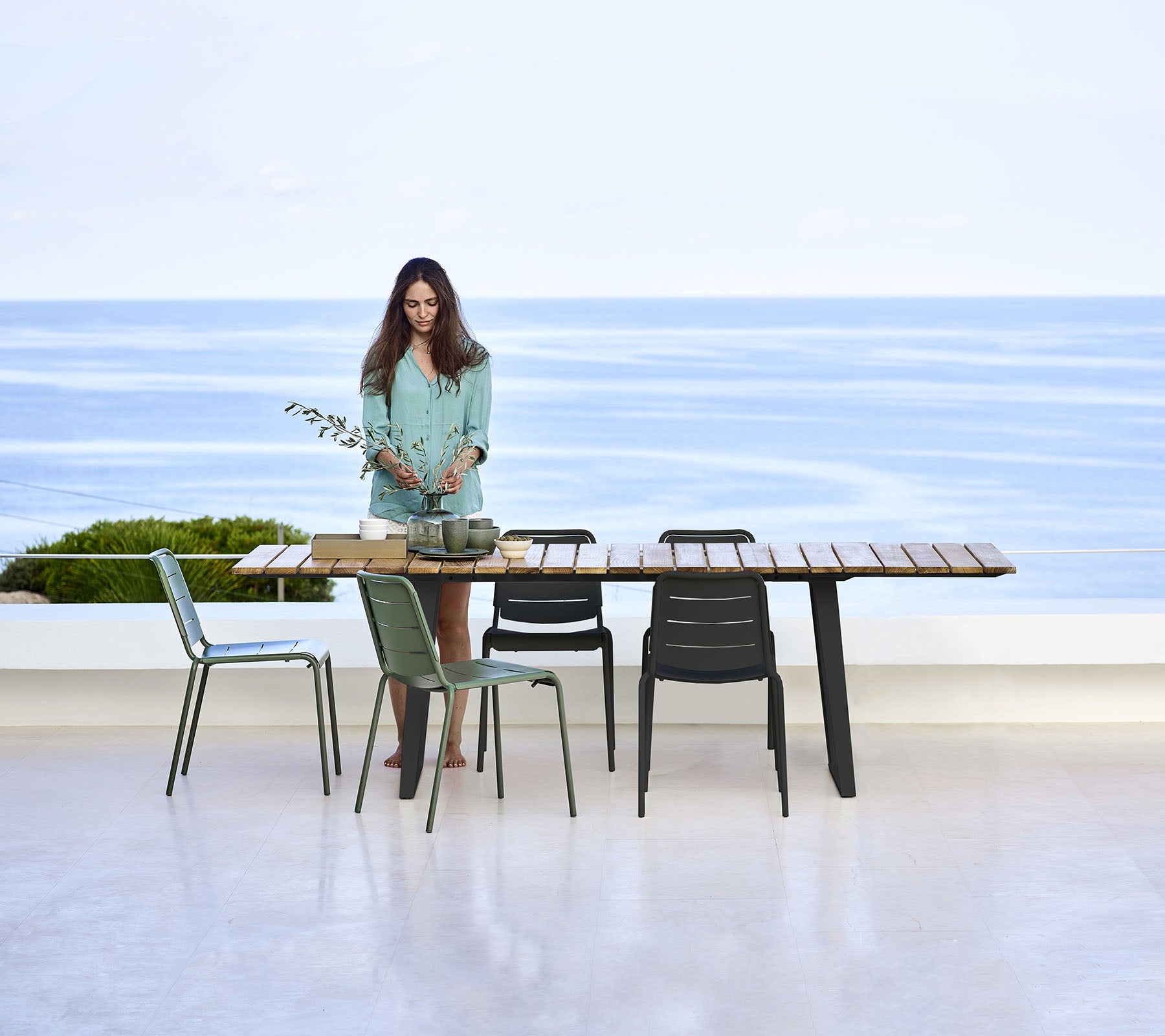 A woman stands at a modern outdoor dining table with chairs, overlooking a serene ocean view.