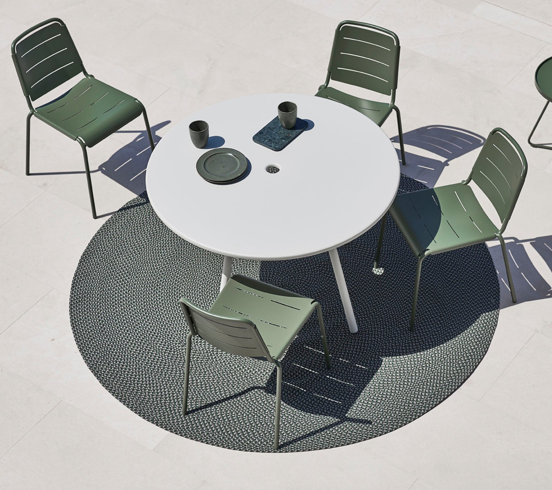 Modern outdoor dining area featuring a round white table, green chairs, and a circular rug. Two cups and a plate are on the table.