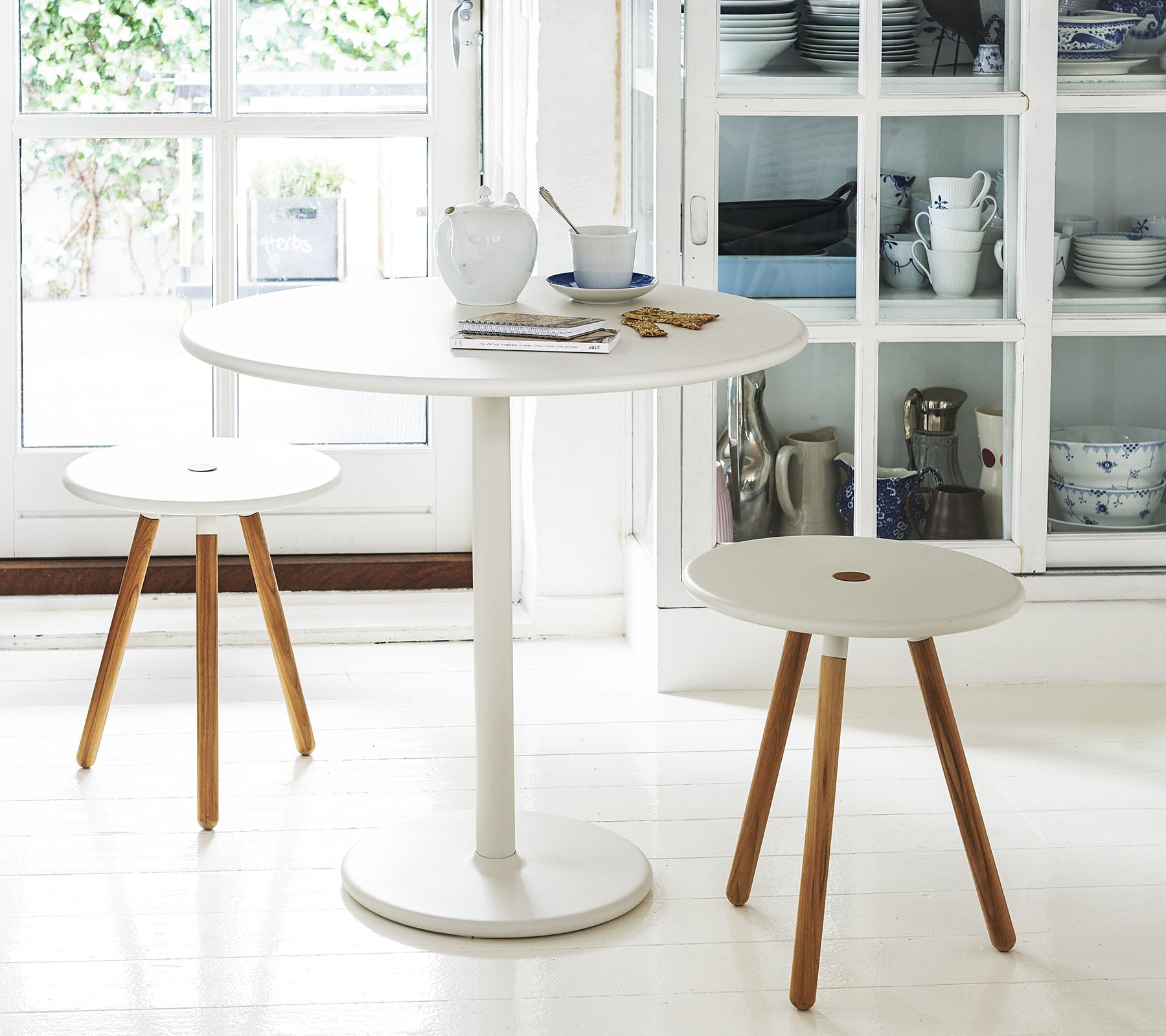 A round white table accompanied by two minimalist stools with wooden legs, set against a backdrop of shelves with kitchenware.