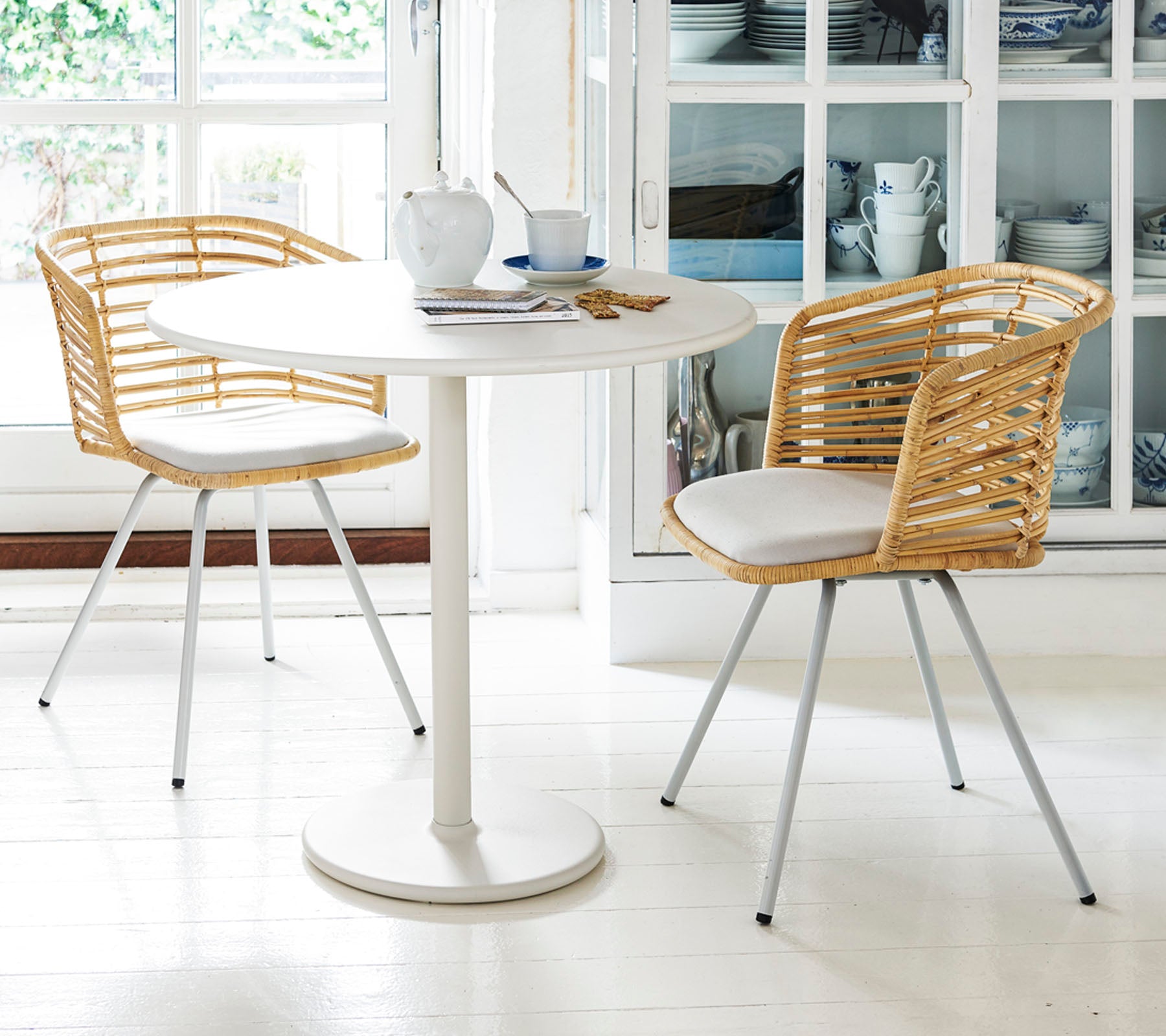 A round table with two stylish chairs features woven backs and cushions, set with teacups and a saucer, creating a cozy dining space.