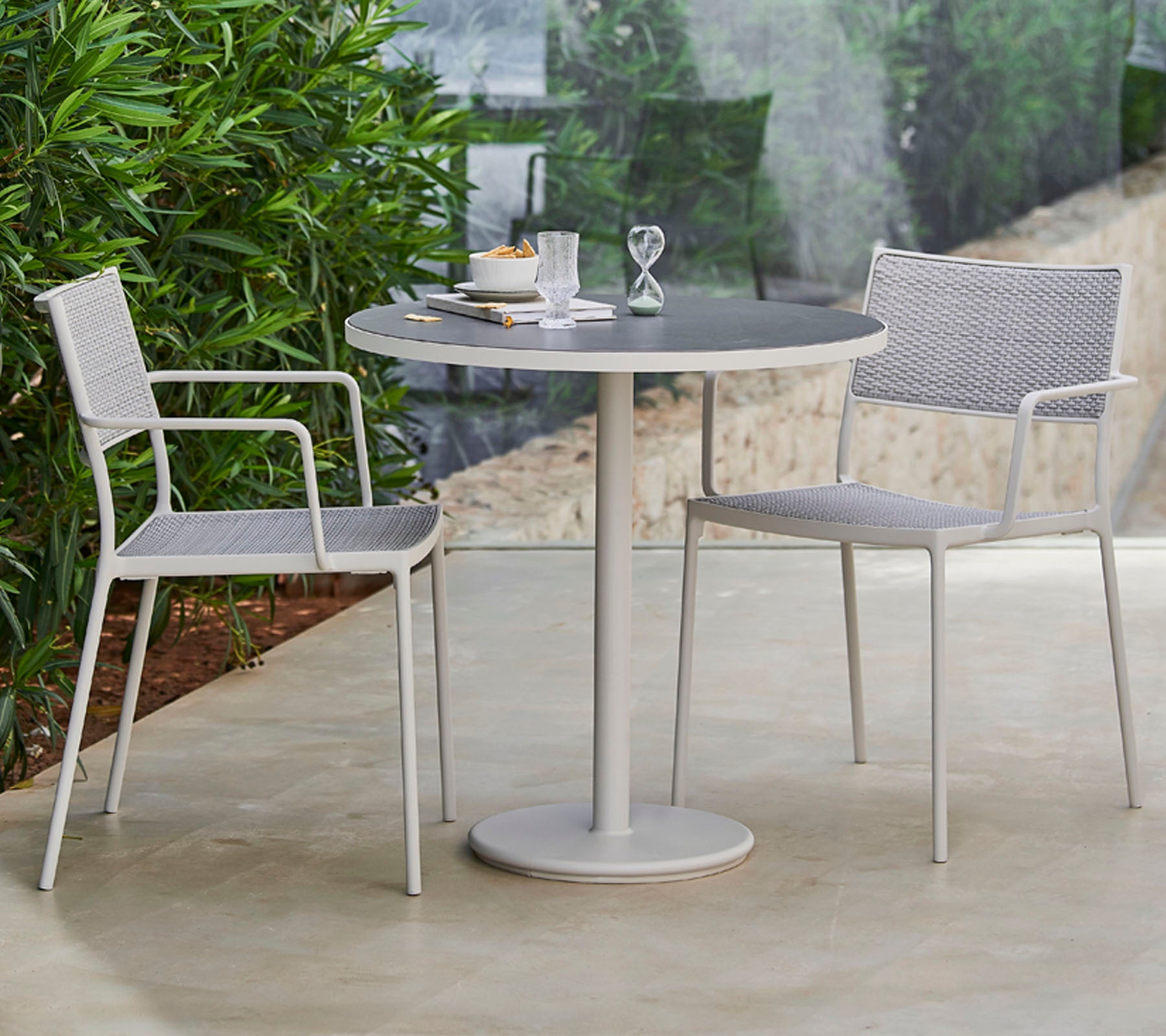 A stylish round table surrounded by two modern chairs, set with tableware and a decorative item, framed by lush greenery.