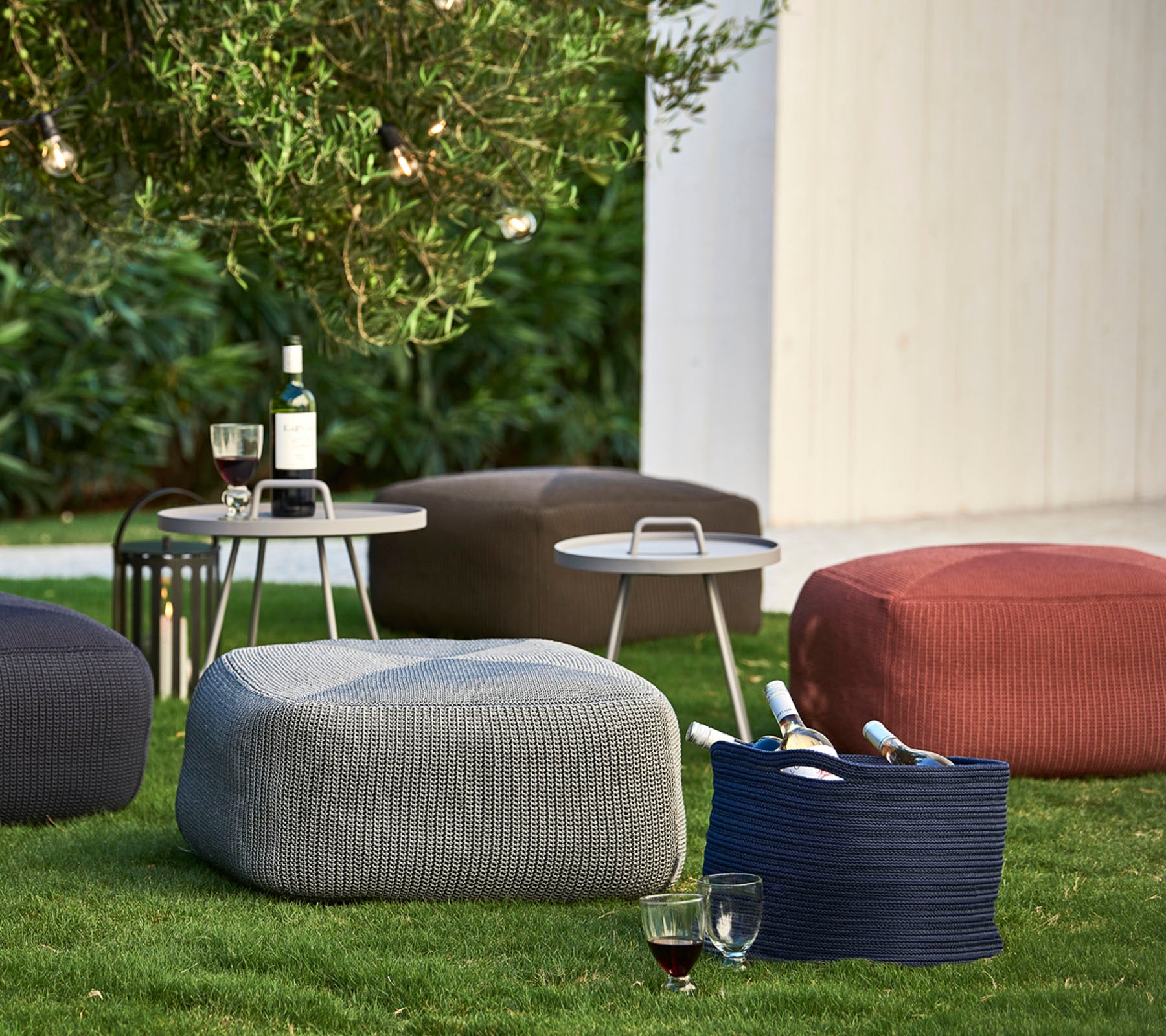 Textured poufs in various colors arranged on grass, accompanied by a small table with drinks and wine bottles.