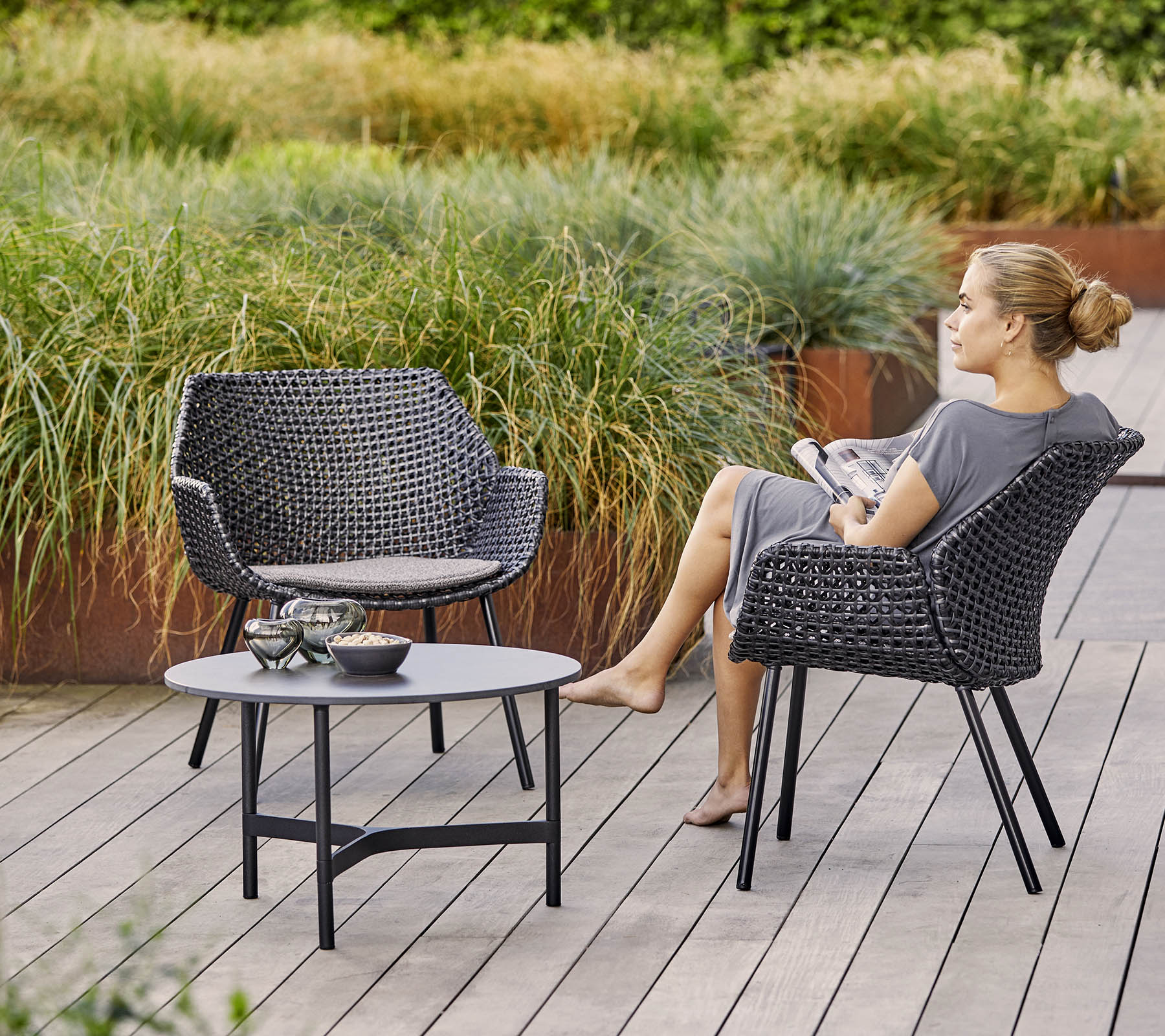 Two stylish woven chairs with a matching table, surrounded by lush greenery. A woman relaxes while holding a magazine.