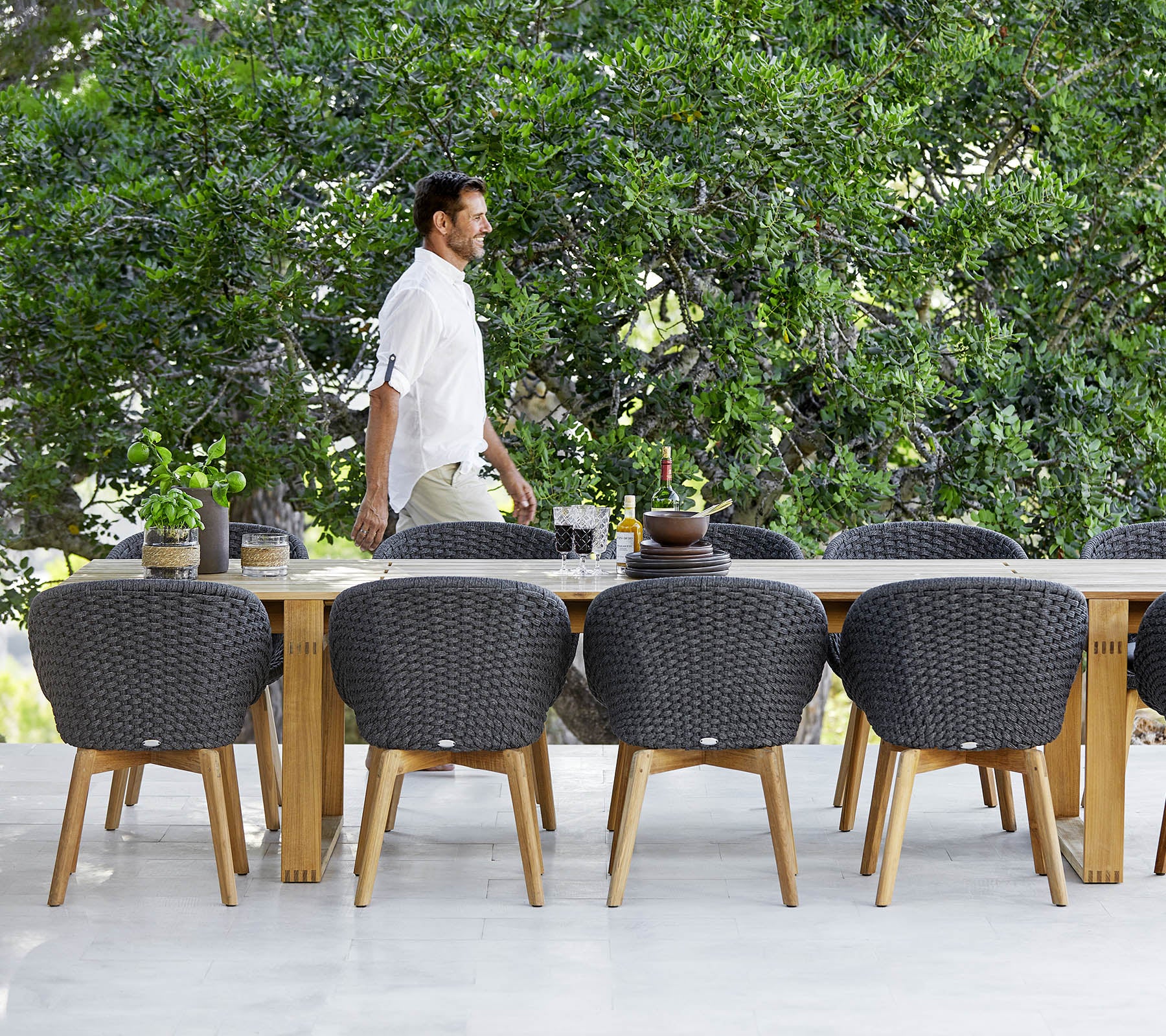 A man walks past a large wooden dining table surrounded by modern, woven chairs and greenery in the background.