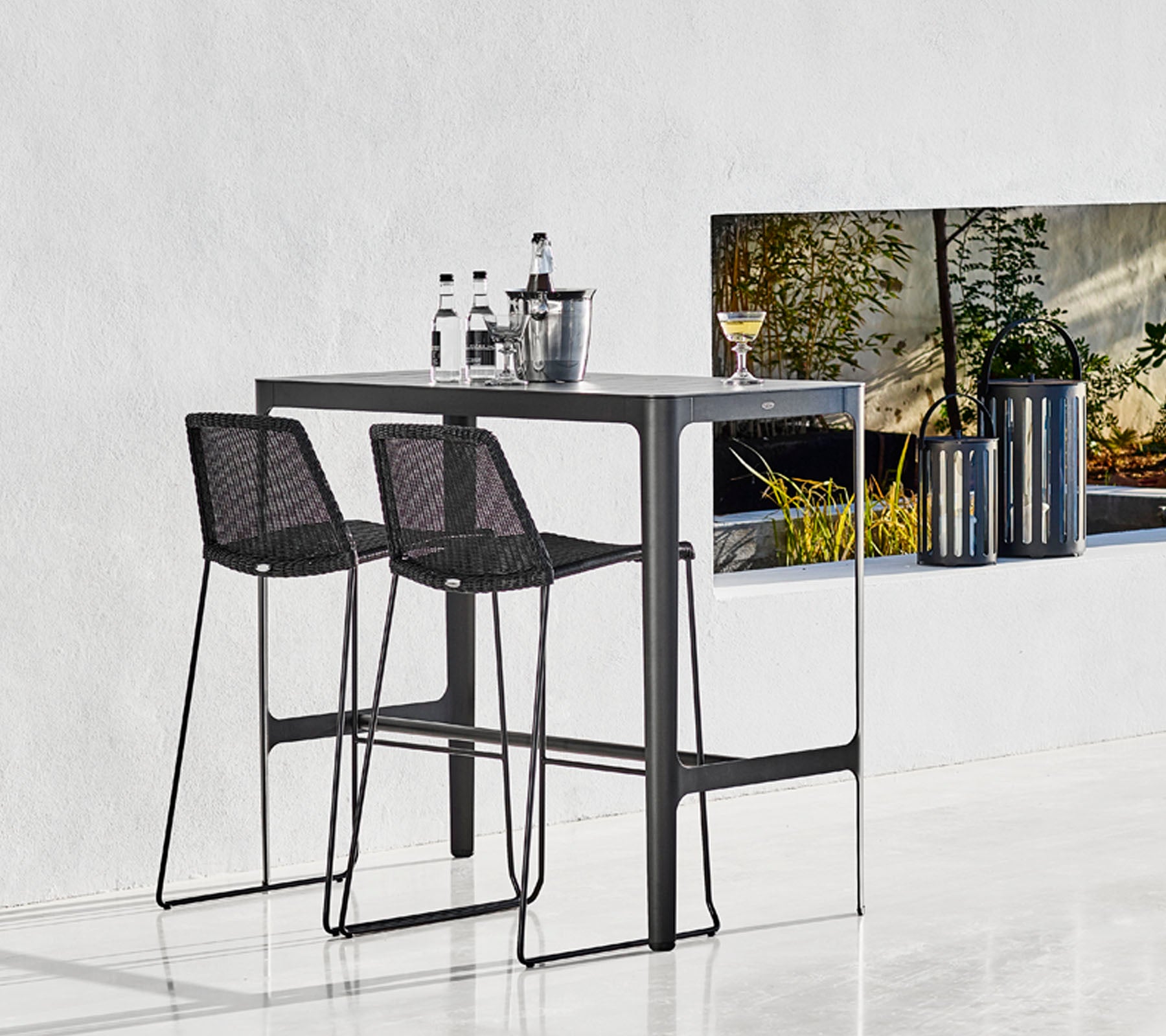 Sleek bar table with two black woven stools, accompanied by glassware and a stylish bucket, set against a minimalist backdrop.