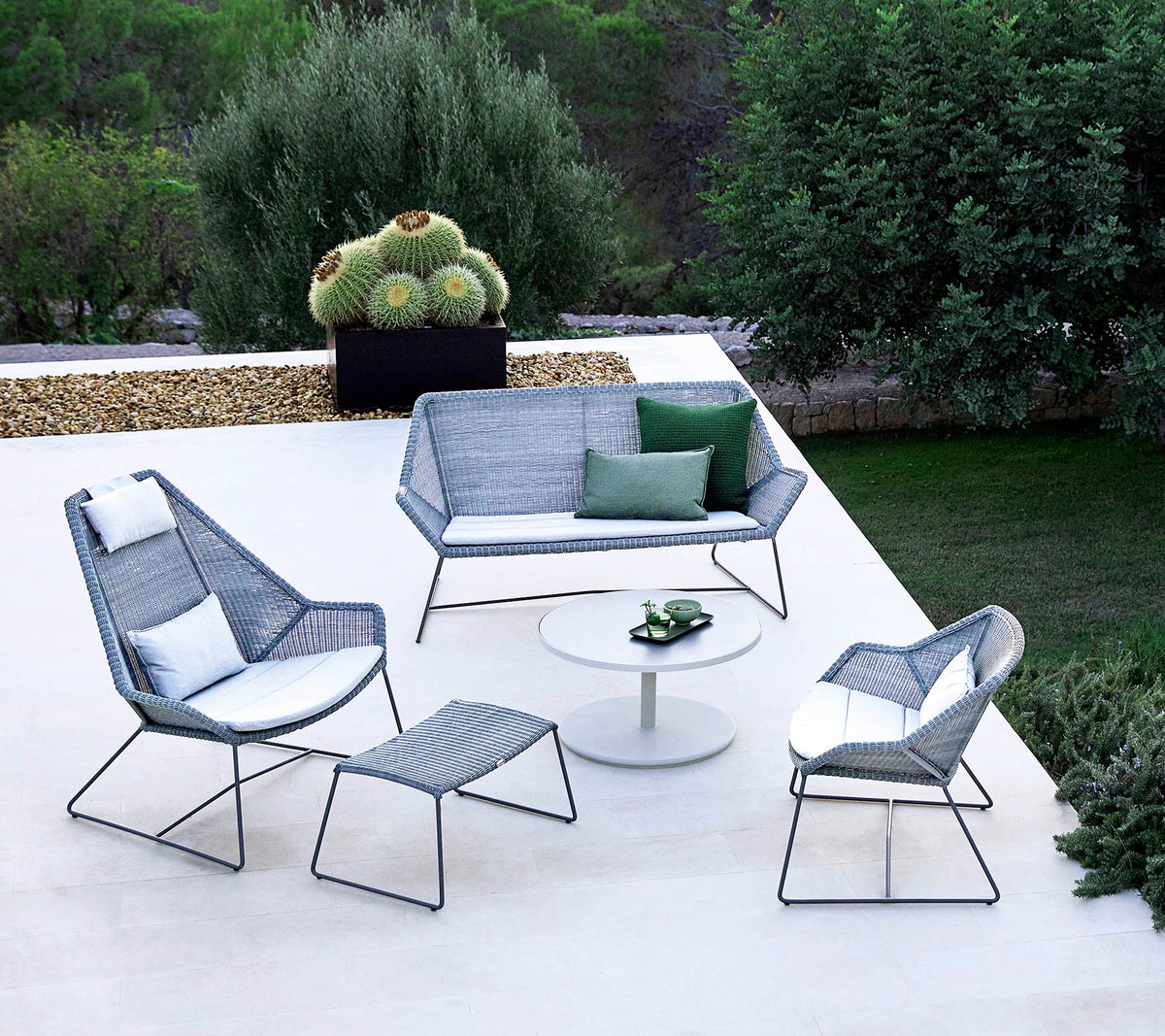 Stylish outdoor seating arrangement featuring a sofa, two armchairs, a footrest, and a coffee table, complemented by greenery in the background.