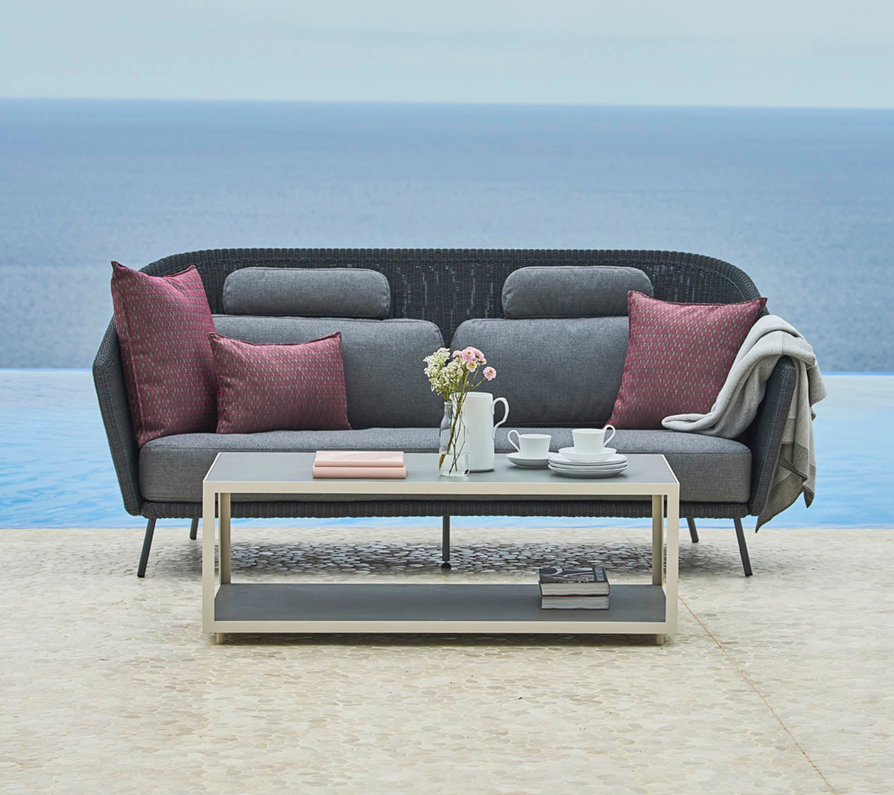 Stylish gray sofa with decorative pillows and a cozy throw, complemented by a sleek coffee table with a teapot and books, set against a serene landscape.