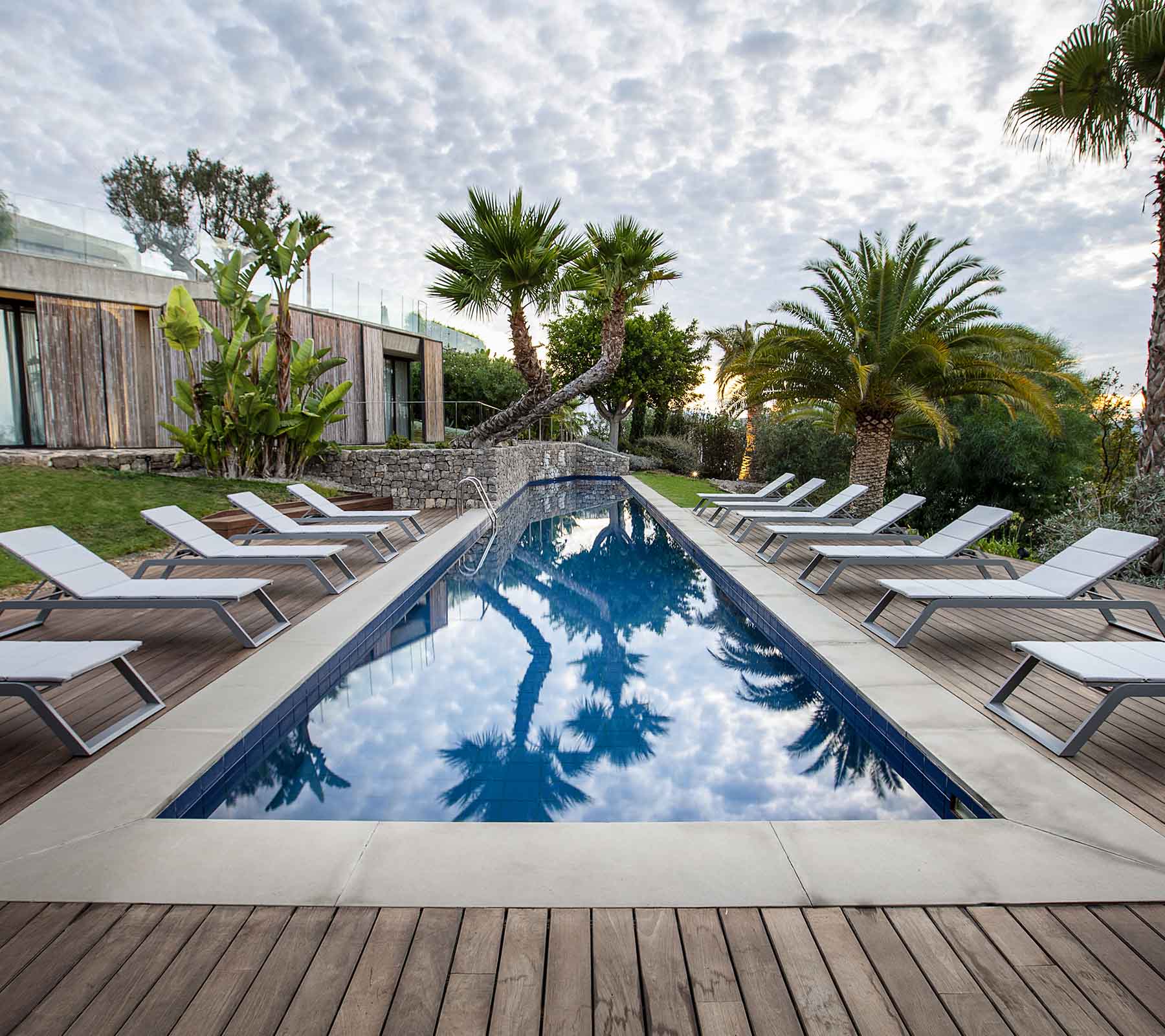 Serene scene featuring a pool surrounded by lounge chairs and palm trees, reflecting the sky and lush greenery.
