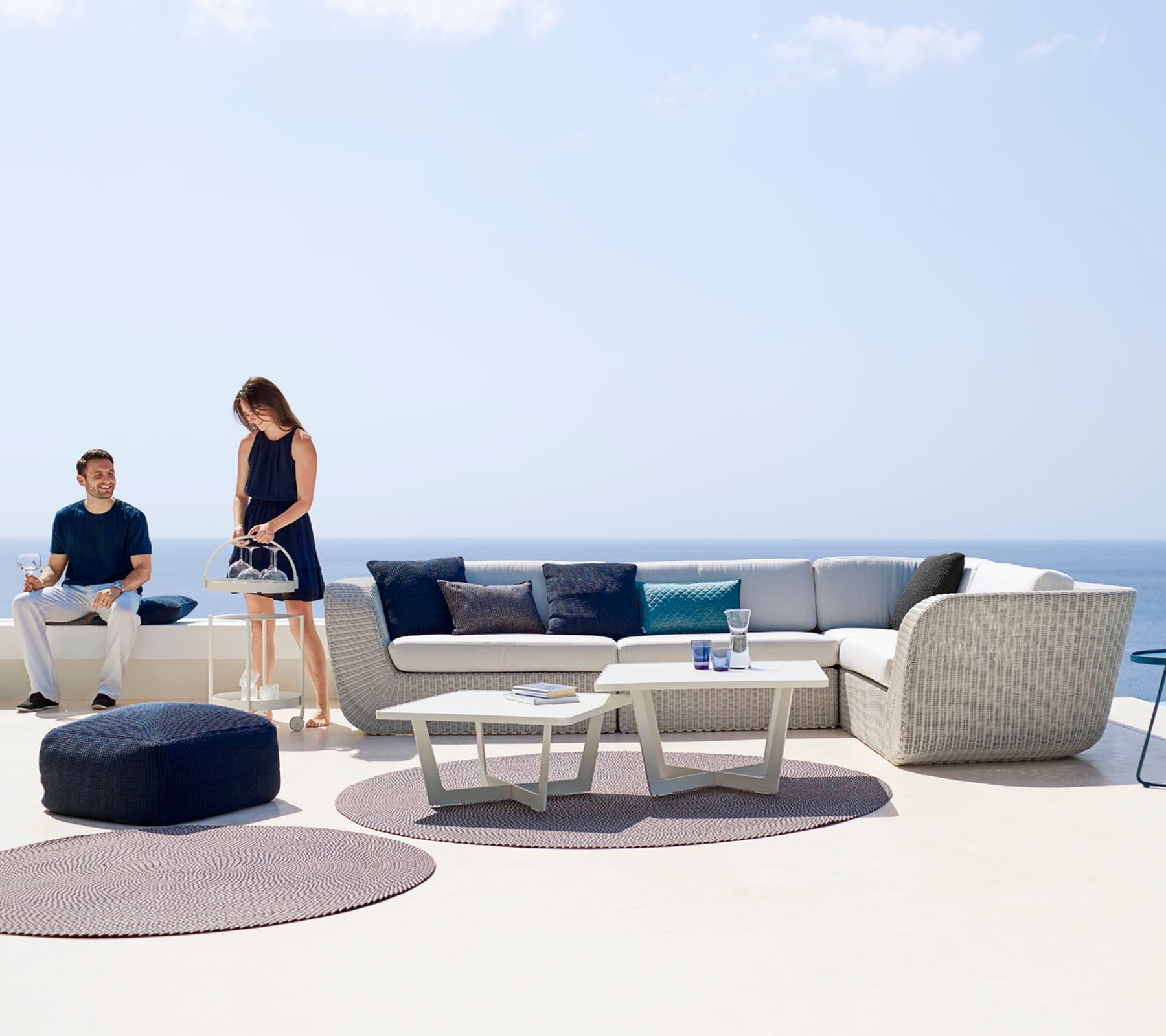 A modern outdoor seating area featuring a sectional sofa, coffee table, and decorative rugs, with two people enjoying the space.