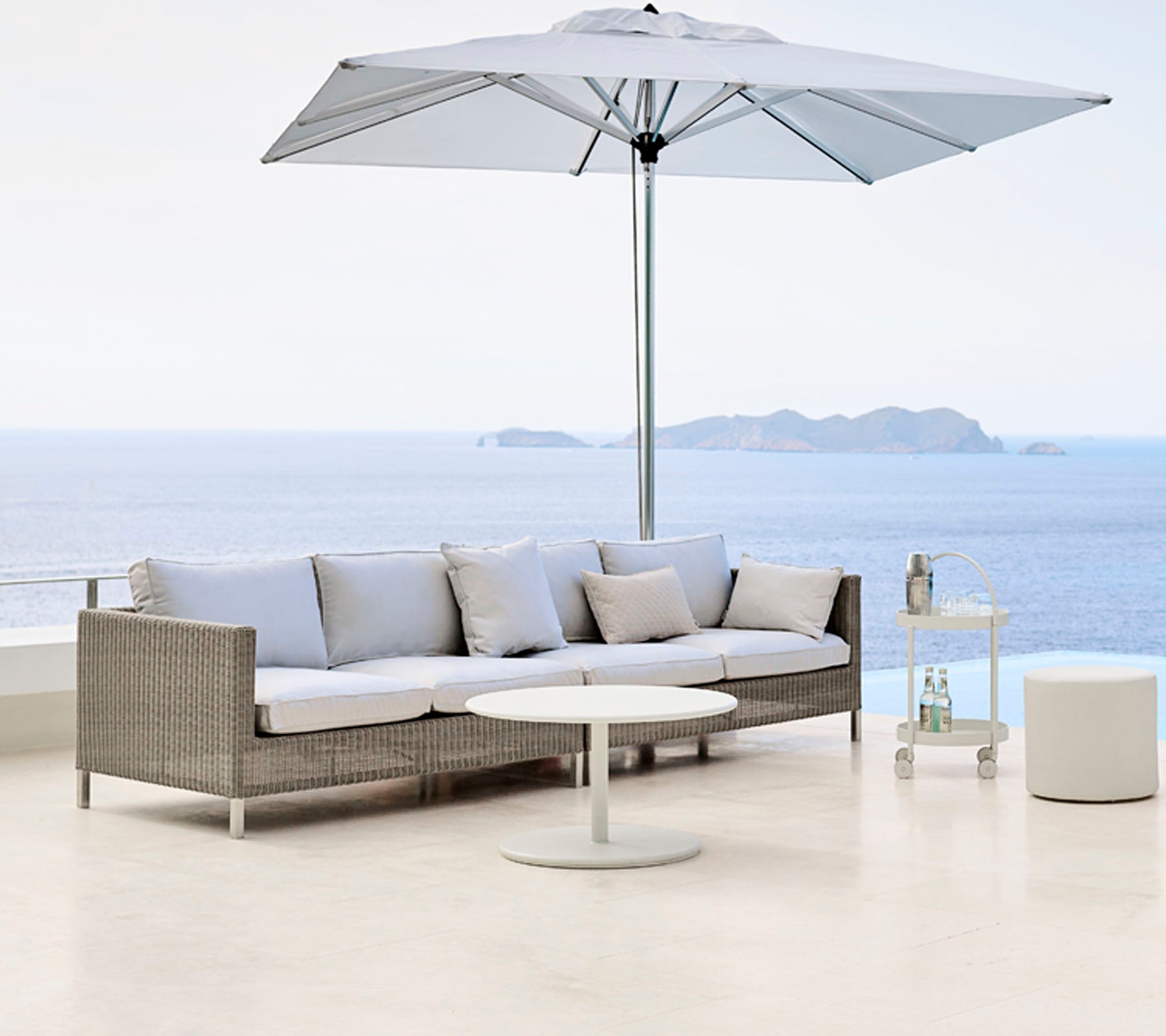 Stylish outdoor seating area with a light gray sofa, round table, and a large umbrella, overlooking a serene body of water.