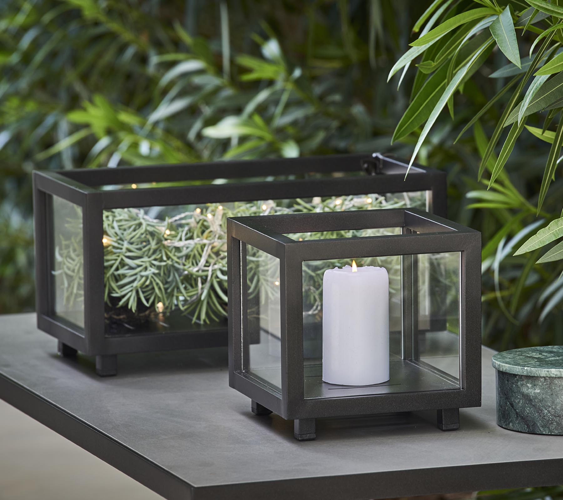 Two stylish lanterns with glass panels, one holding a white candle and the other featuring greenery, placed on a table amid lush foliage.