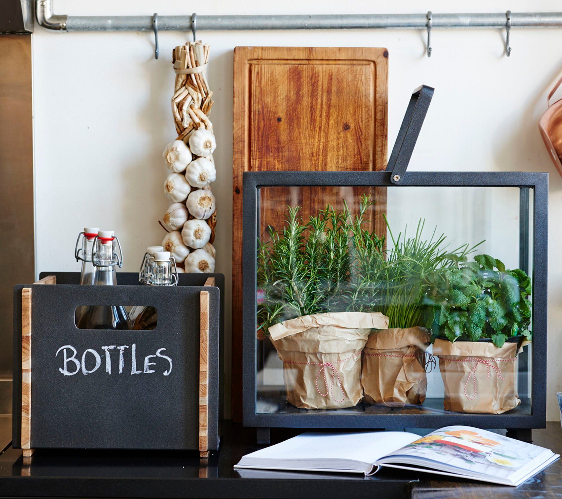 A stylish display featuring a black box labeled "BOTTLES," a glass container with potted herbs, and a cookbook on a surface.