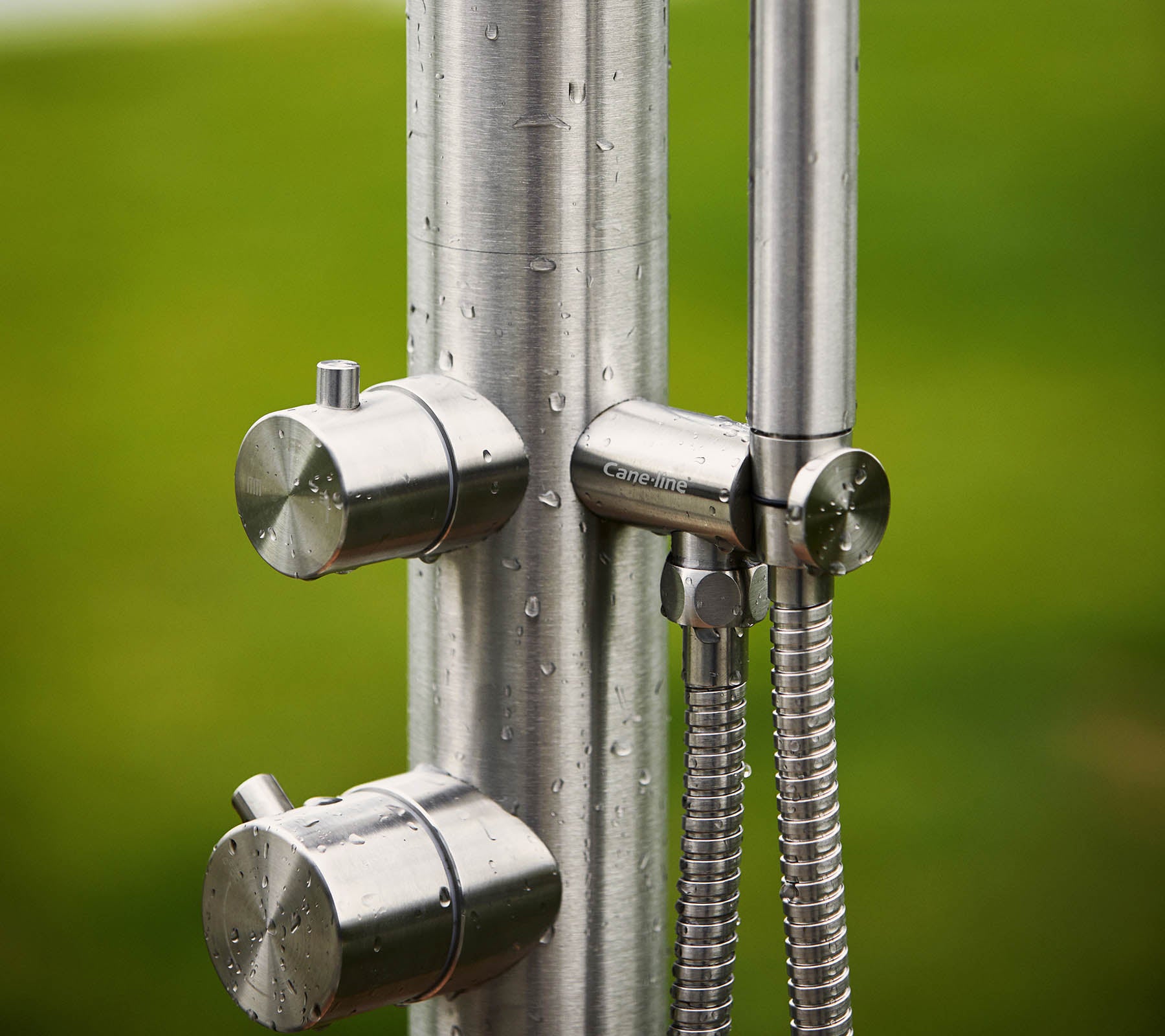 Stainless steel shower unit featuring two knobs for temperature control and a handheld showerhead, set against a green background.
