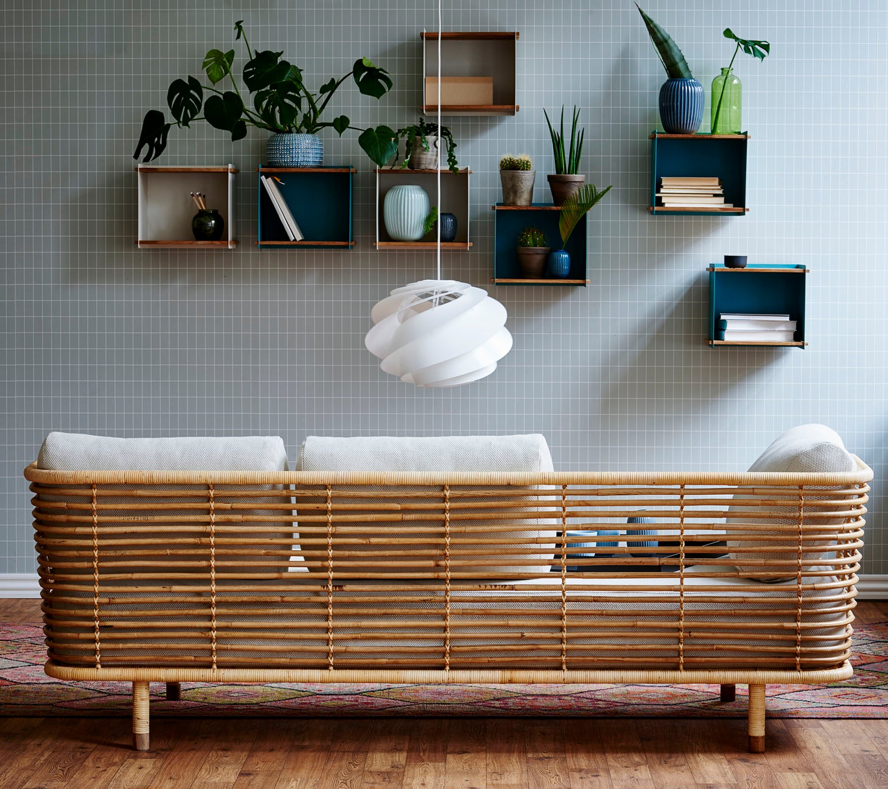 A stylish rattan sofa with light cushions against a textured wall, complemented by wall-mounted shelves displaying plants and decorative items.