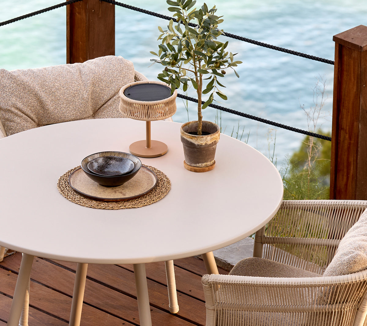 White round table with woven chairs, decorative plant, and a black bowl in a modern lounge setting.