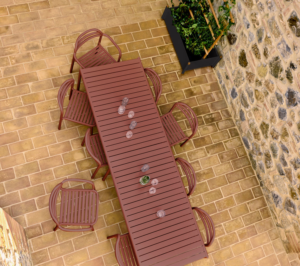 A desert red Bliss table with matching chairs arranged around it, set on a stone floor with a planter in the background.