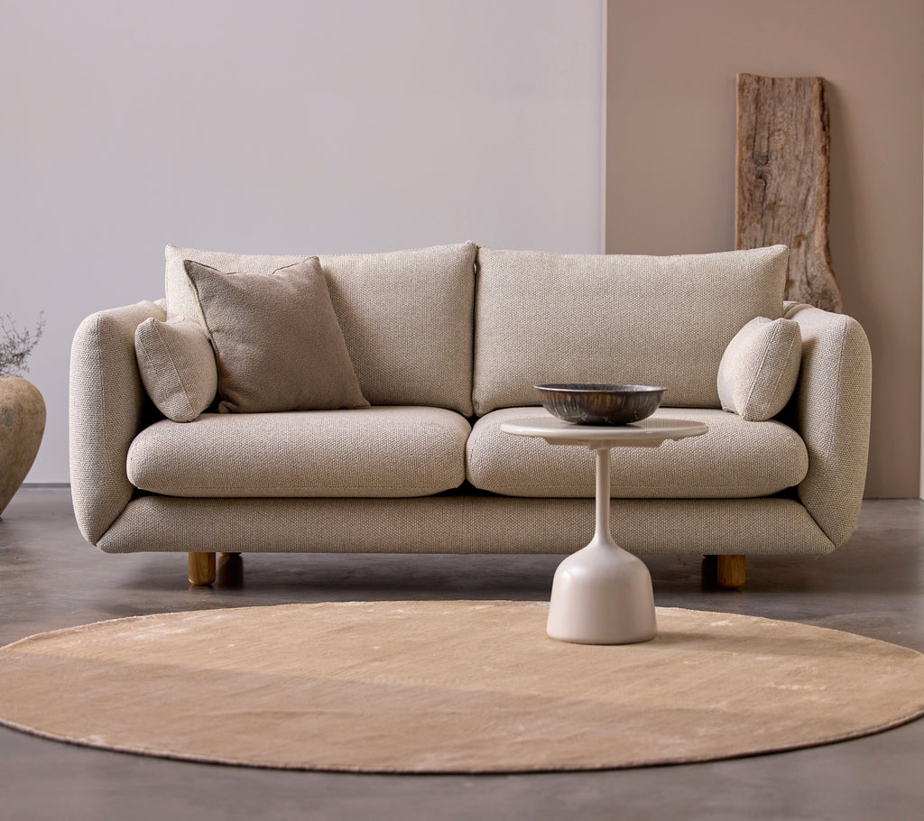Bloom 2-seater sofa in desert sand with rounded cushions and teak legs, paired with a minimalistic side table on a circular rug.