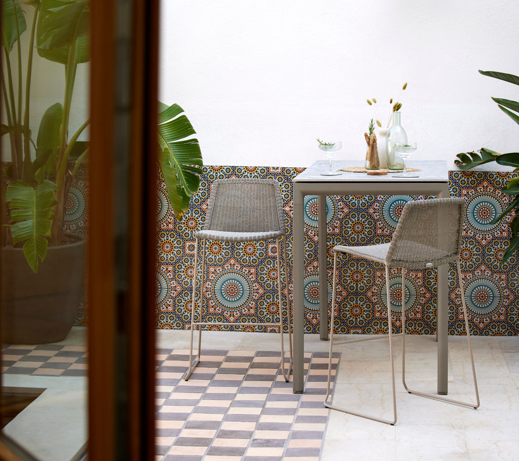 Breeze bar chairs in taupe with a sleek drop bar table, surrounded by decorative tiled walls and lush plants.