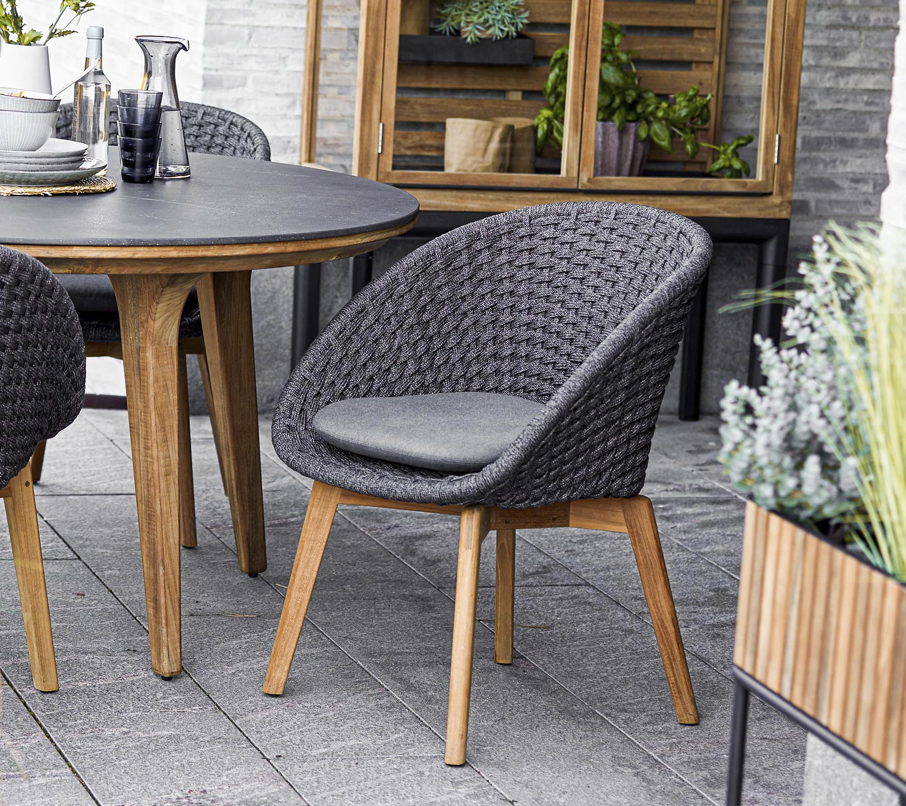 Stylish woven chair with a padded seat, paired with a round table, and surrounded by greenery in planters. Perfect for social gatherings.