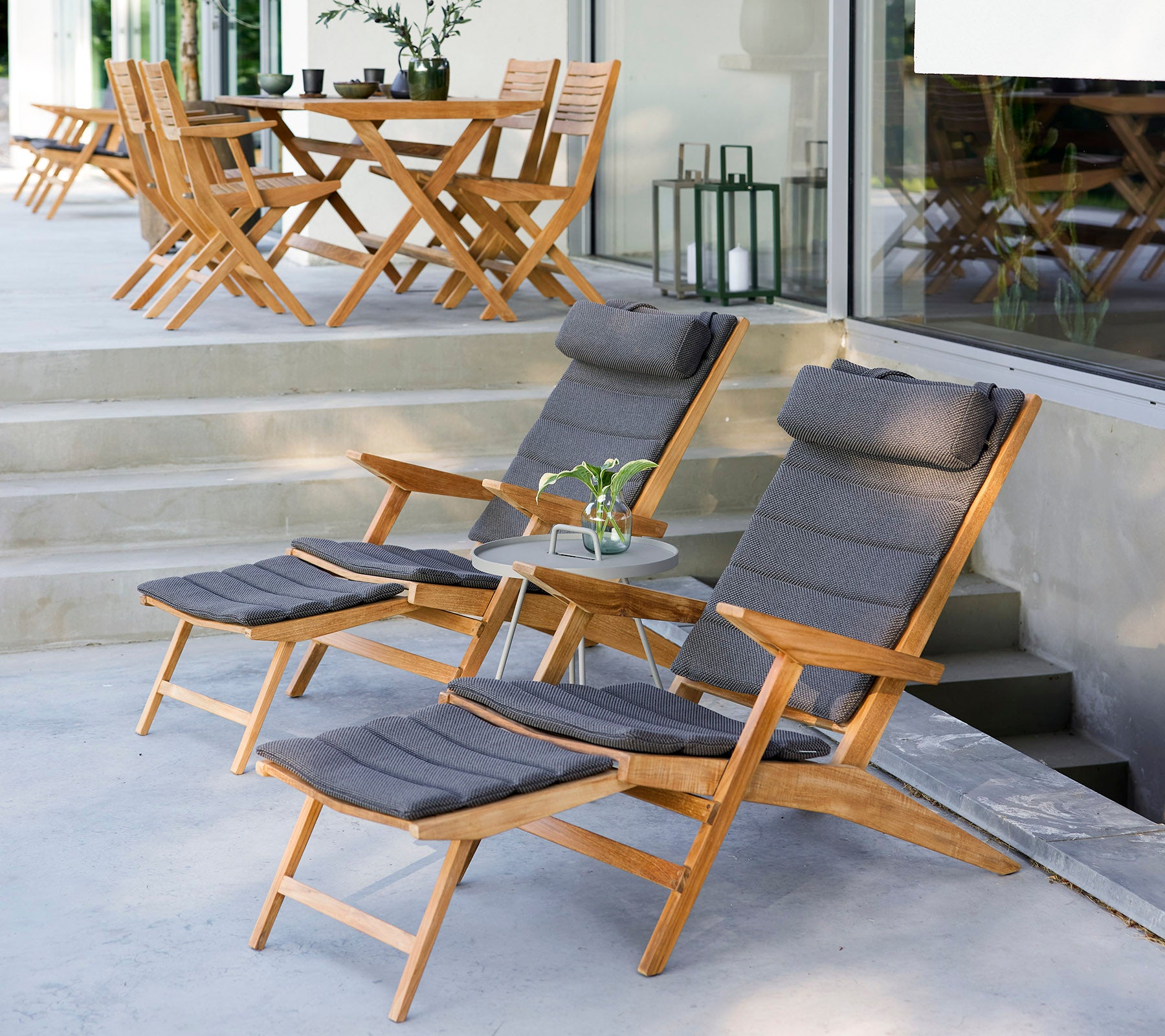 Two wooden lounge chairs with cushions, a small side table, and greenery in a stylish outdoor setting with additional chairs in the background.