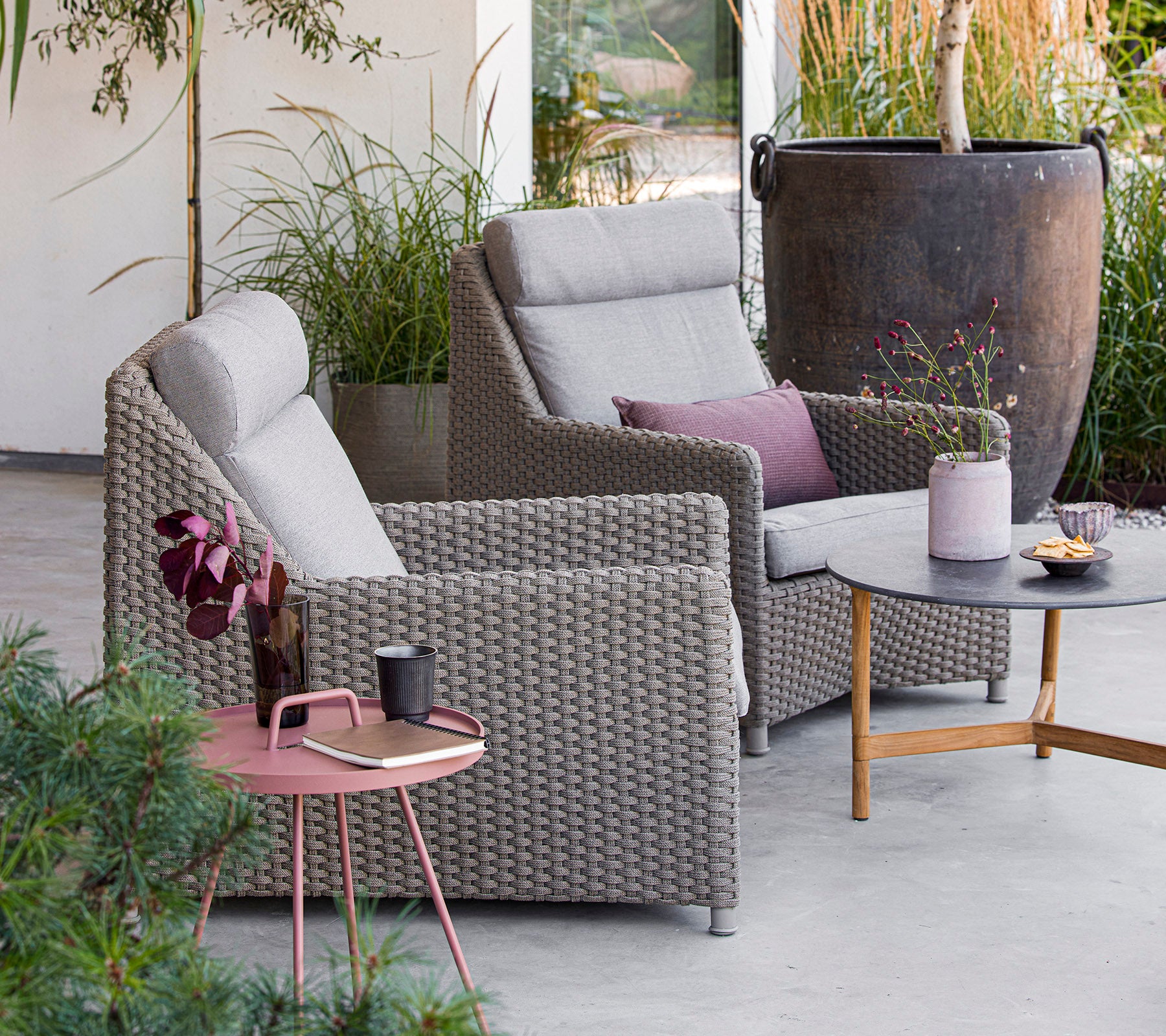 Two comfortable grey chairs with cushions, a round table with snacks and drinks, surrounded by greenery and decor. Perfect for relaxation.