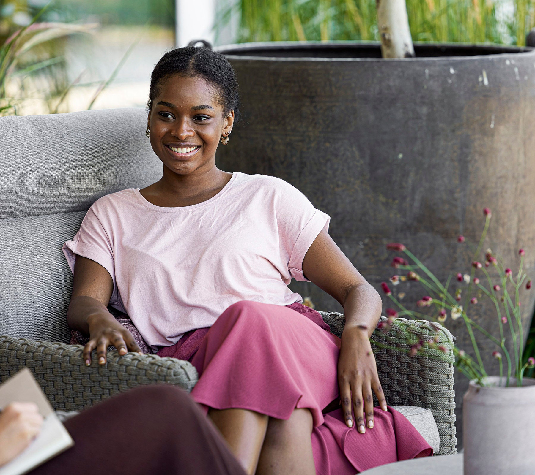 A smiling woman in a pink top and skirt sits comfortably on a couch, enjoying a relaxed moment in a serene setting.
