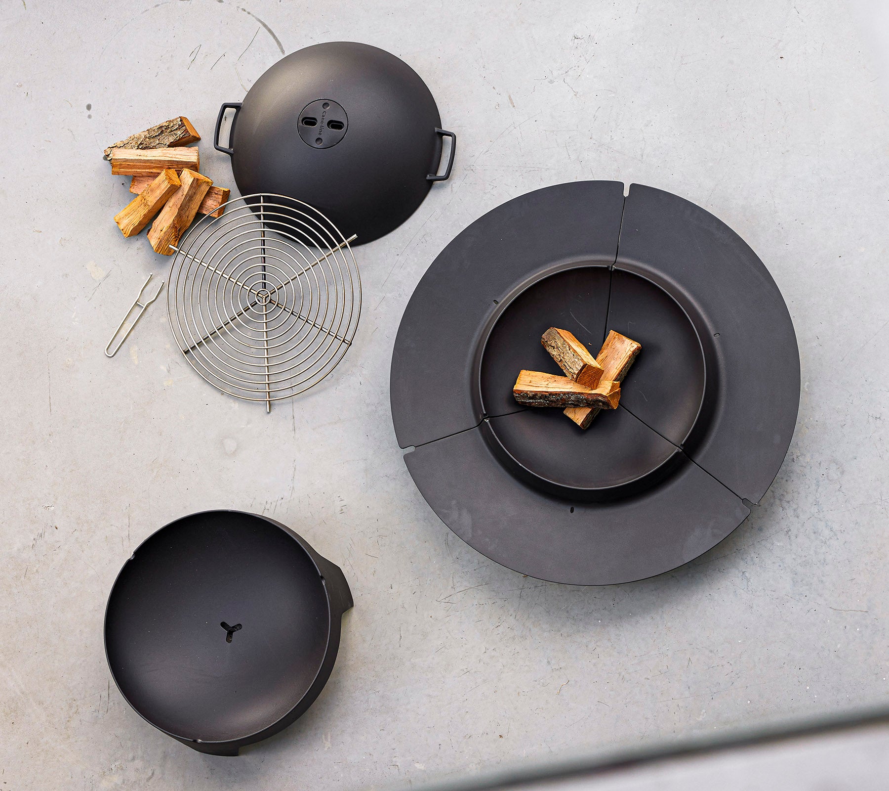Black cooking equipment laid out with firewood and a wire grilling rack, designed for outdoor grilling and fire cooking.