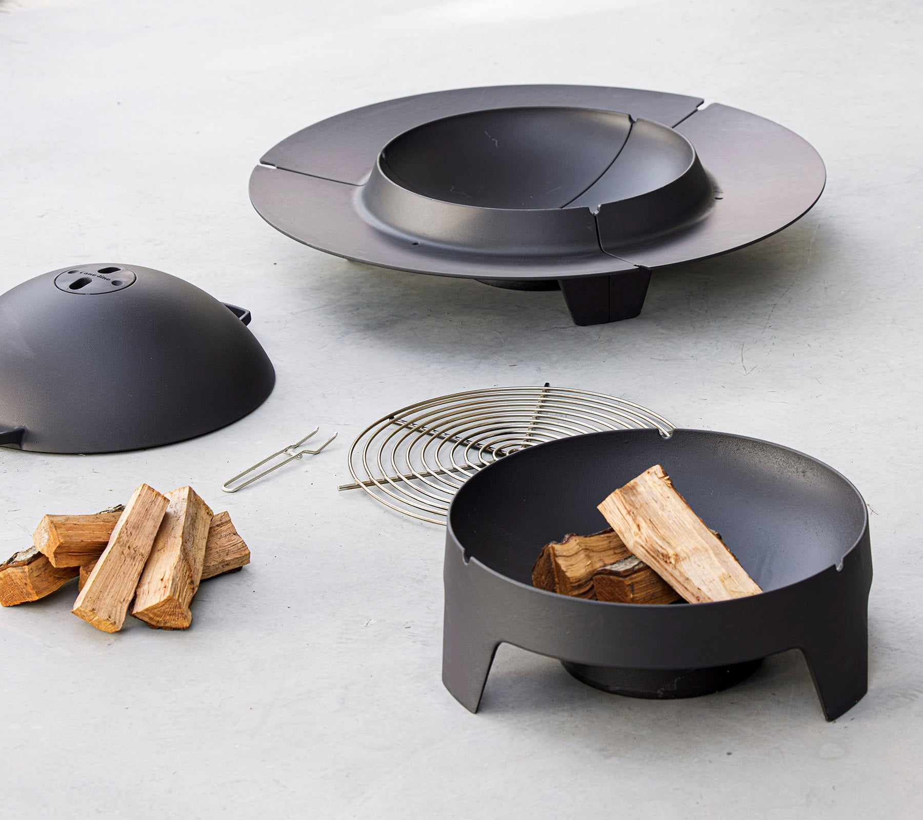 A black fire pit set featuring a bowl, lid, a grate, and stacked firewood, designed for outdoor gatherings.