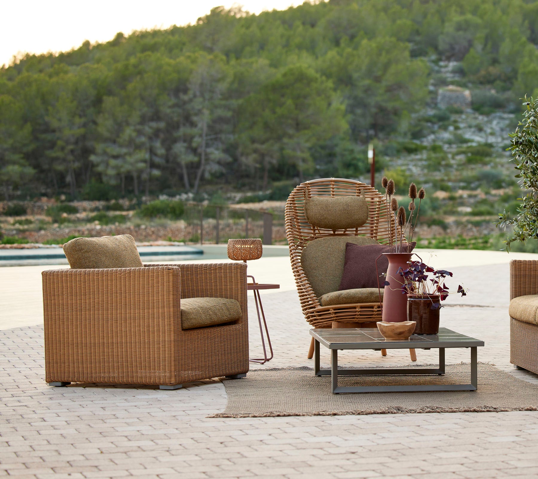 A cozy seating arrangement featuring woven chairs, a side table, and a decorative vase, surrounded by greenery and natural scenery.