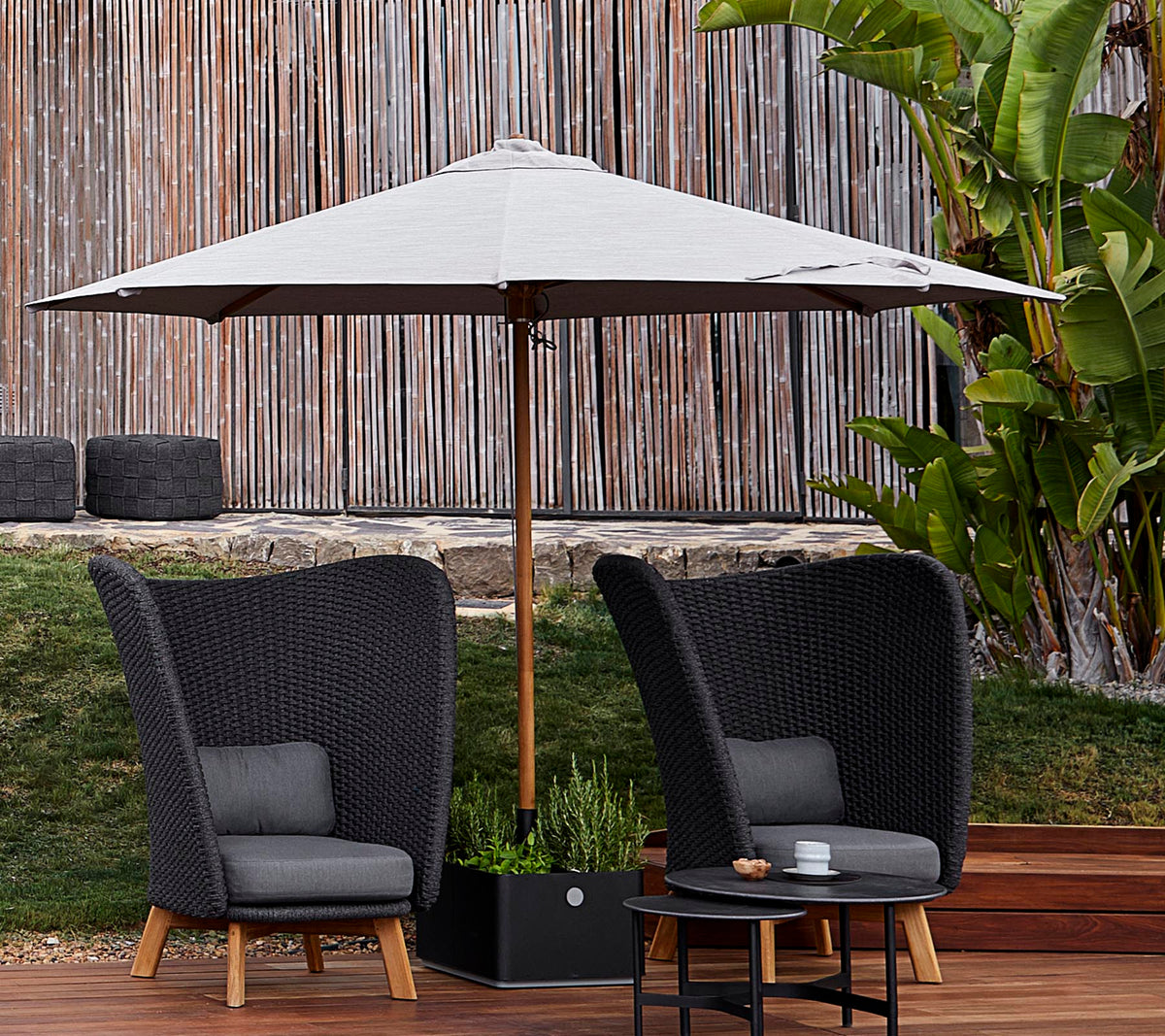 Two stylish woven chairs with cushions sit under a large umbrella, accompanied by a small table, set against a lush green backdrop.