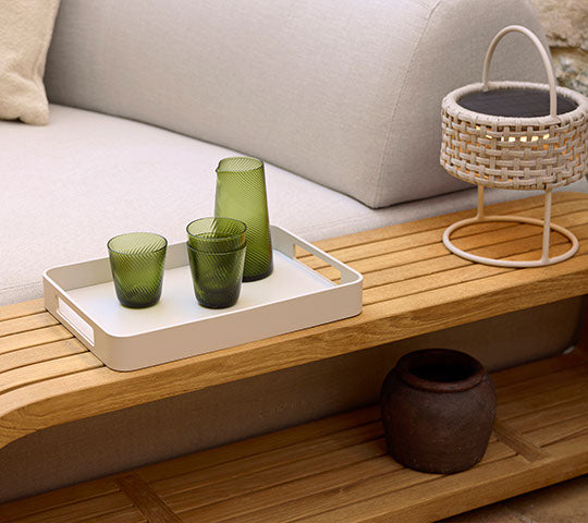 A wooden coffee table featuring a tray with green glassware, a decorative basket, and a dark ceramic pot.