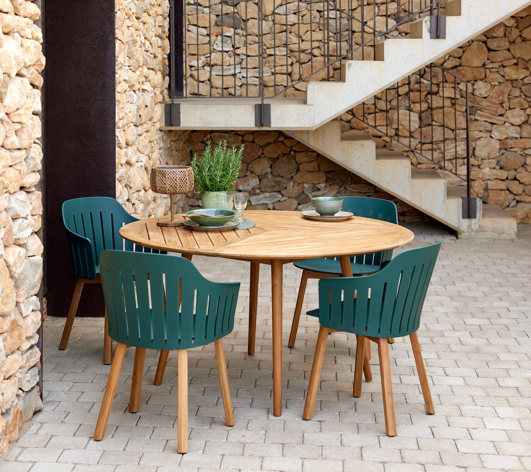 A round wooden table surrounded by four green slatted chairs, complemented by decorative bowls and a small plant.