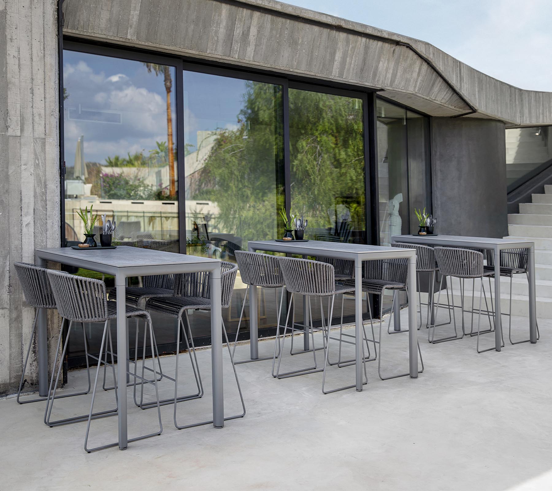 Modern seating area featuring gray tables and chairs, with large windows reflecting greenery outside. Minimalist design aesthetic.
