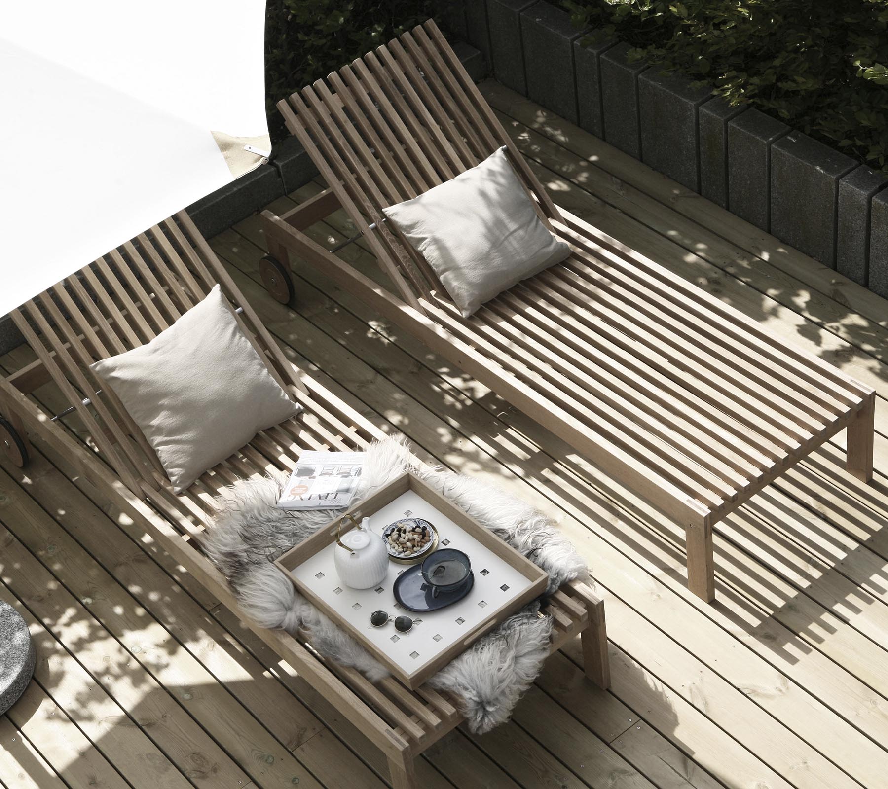 Two wooden lounge chairs with cushions are arranged beside a small table adorned with a cozy throw and decorative items. Natural light filters through.