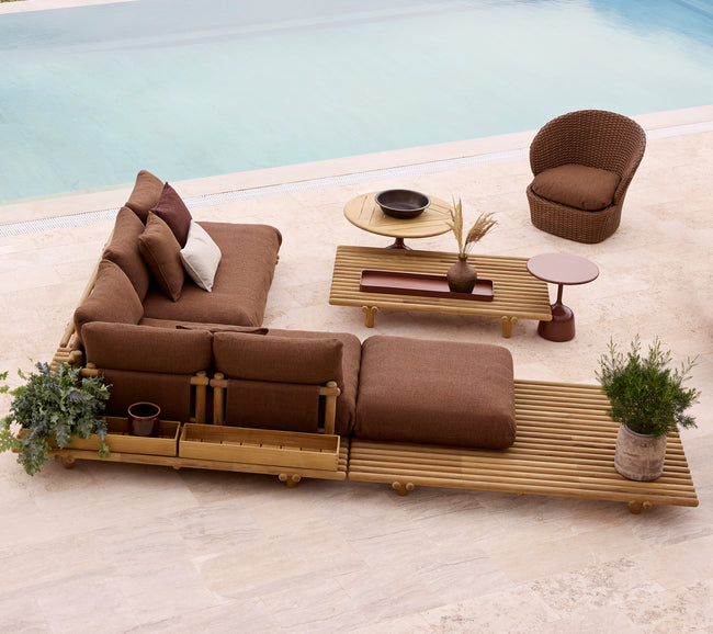 Large corner sofa with brown cushions on a teak platform, accompanied by a lounge chair and modern side tables, surrounded by greenery.