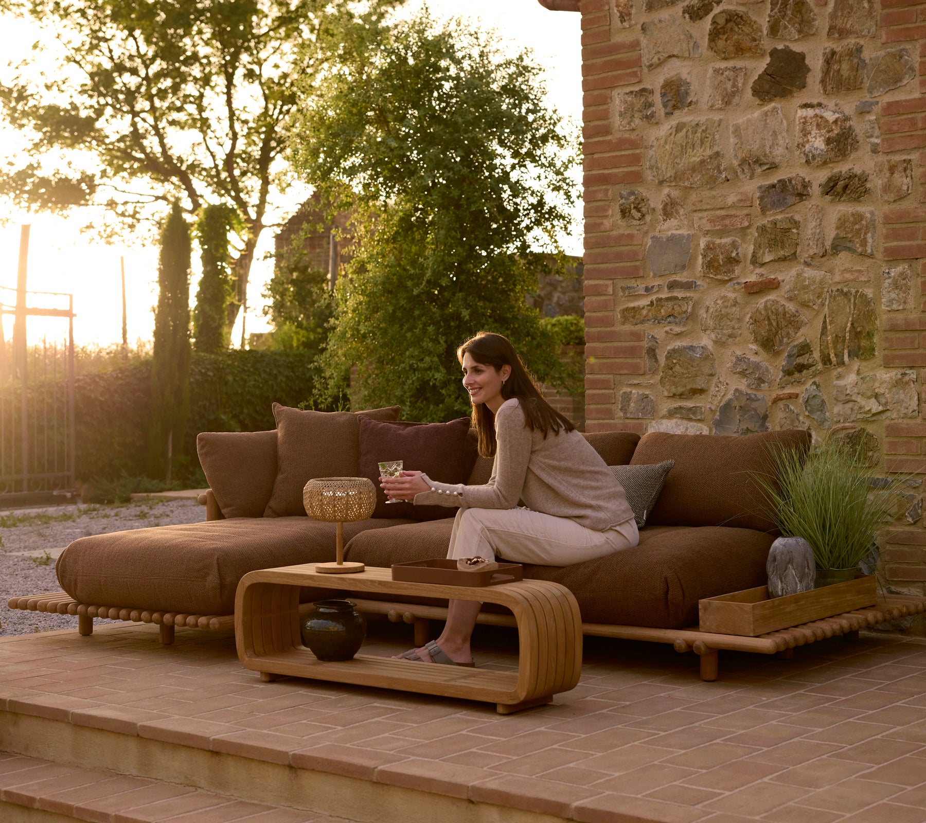 A woman relaxes on a contemporary teak outdoor sofa with a mellow coffee table and decorative tray, set against a stone backdrop.